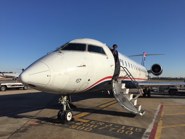 Person boarding a white private jet with stairs on tarmac, clear blue sky, airport vehicles in background.