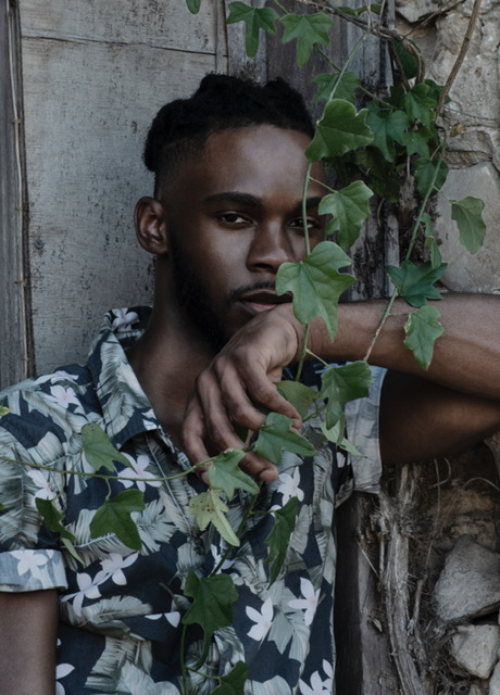Young person with short hair and patterned shirt, partially obscured by green ivy leaves, standing against a concrete wall.
