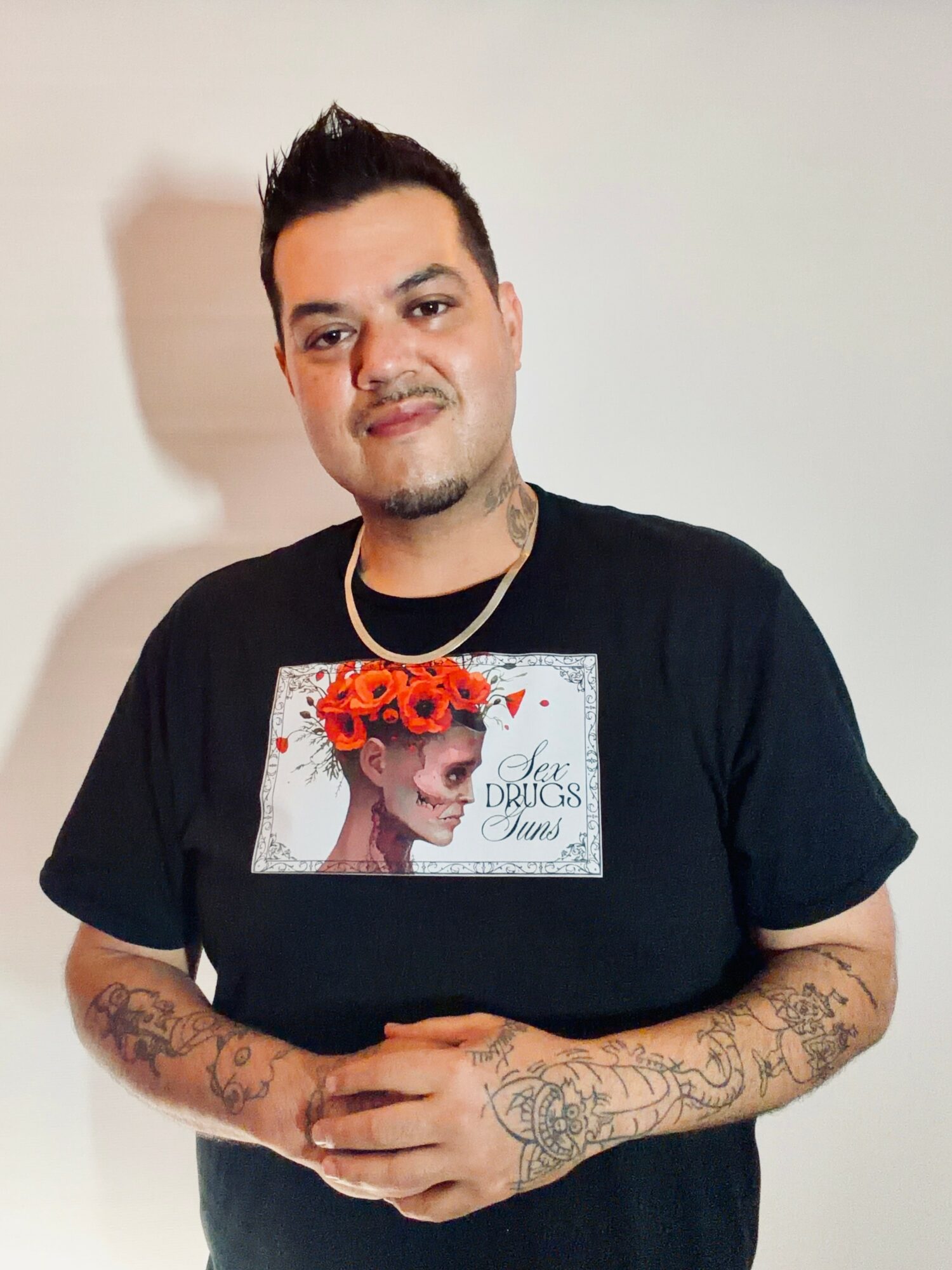 Young man with short dark hair and tattoos on arms, wearing a black T-shirt with a graphic of a woman with flowers in her hair, standing against a plain background.