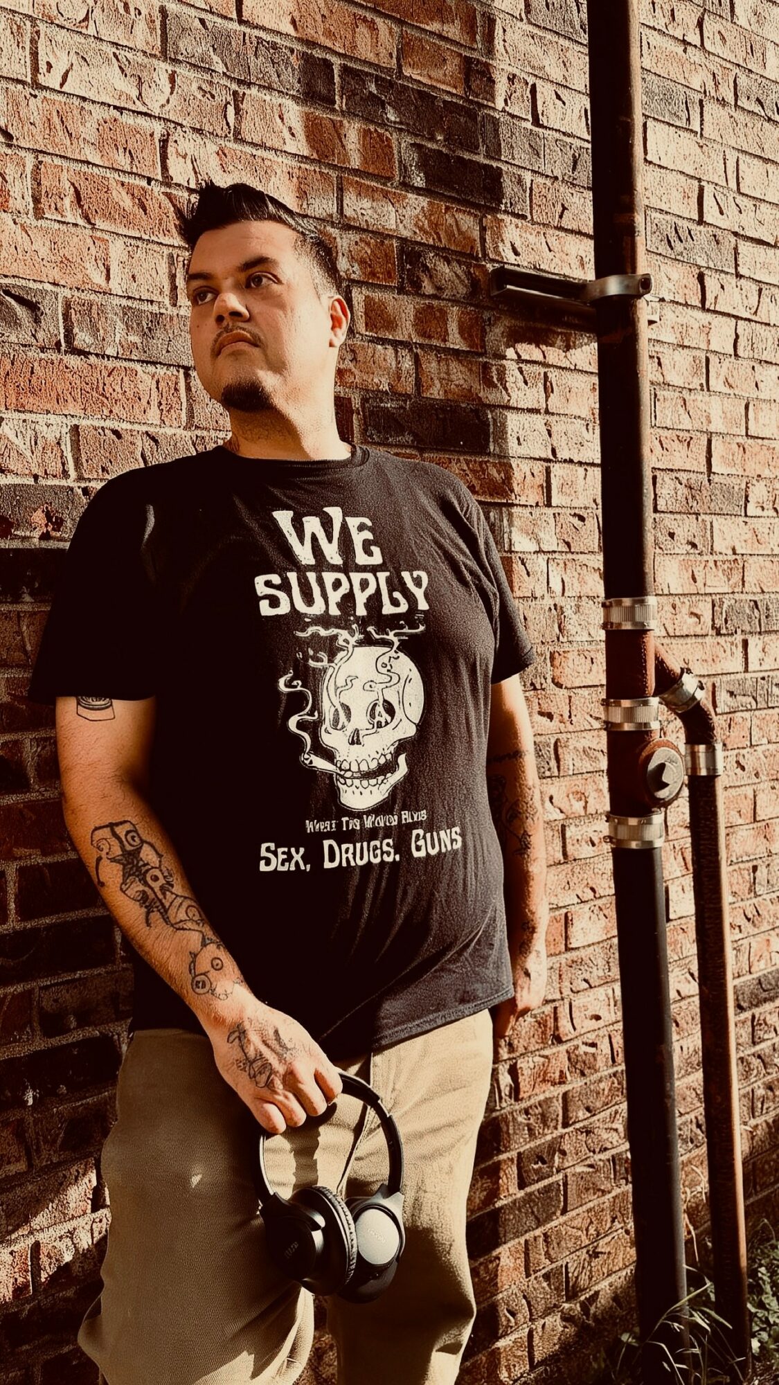 Young man with tattoos on his left arm, wearing a black T-shirt with skull graphic and text, standing against a brick wall.