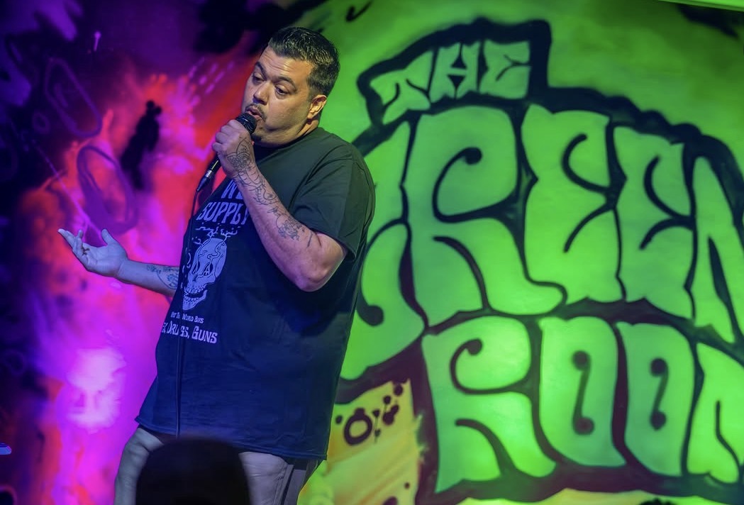 Performer with tattoos singing into a microphone on stage with colorful graffiti background.