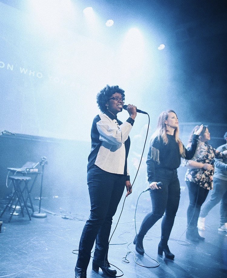 Three people singing on stage with blue lighting, one holding a microphone, others with microphones, in a concert setting.