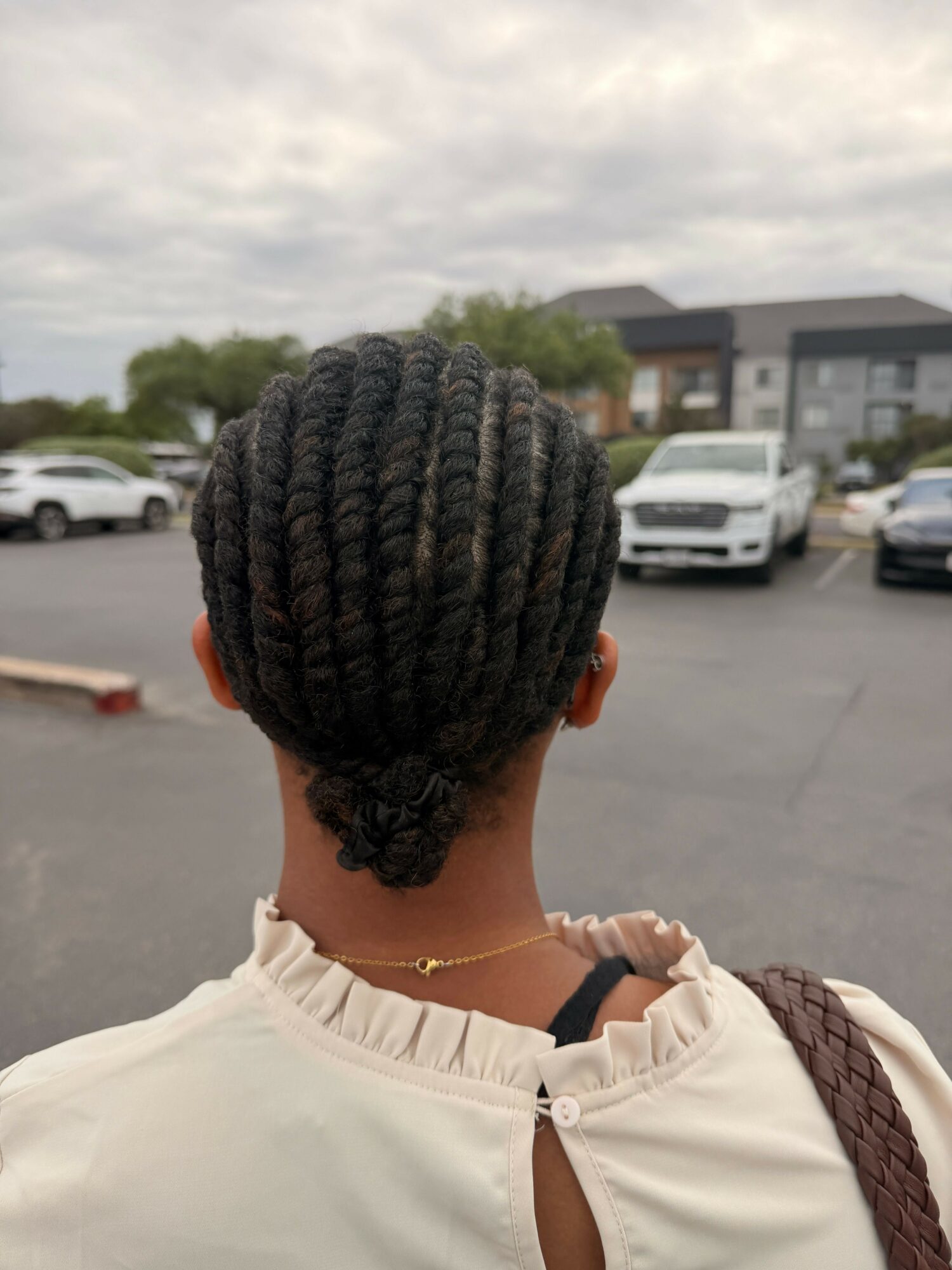 Back view of a person with braided hair wearing a light-colored top, standing outdoors near parked cars.