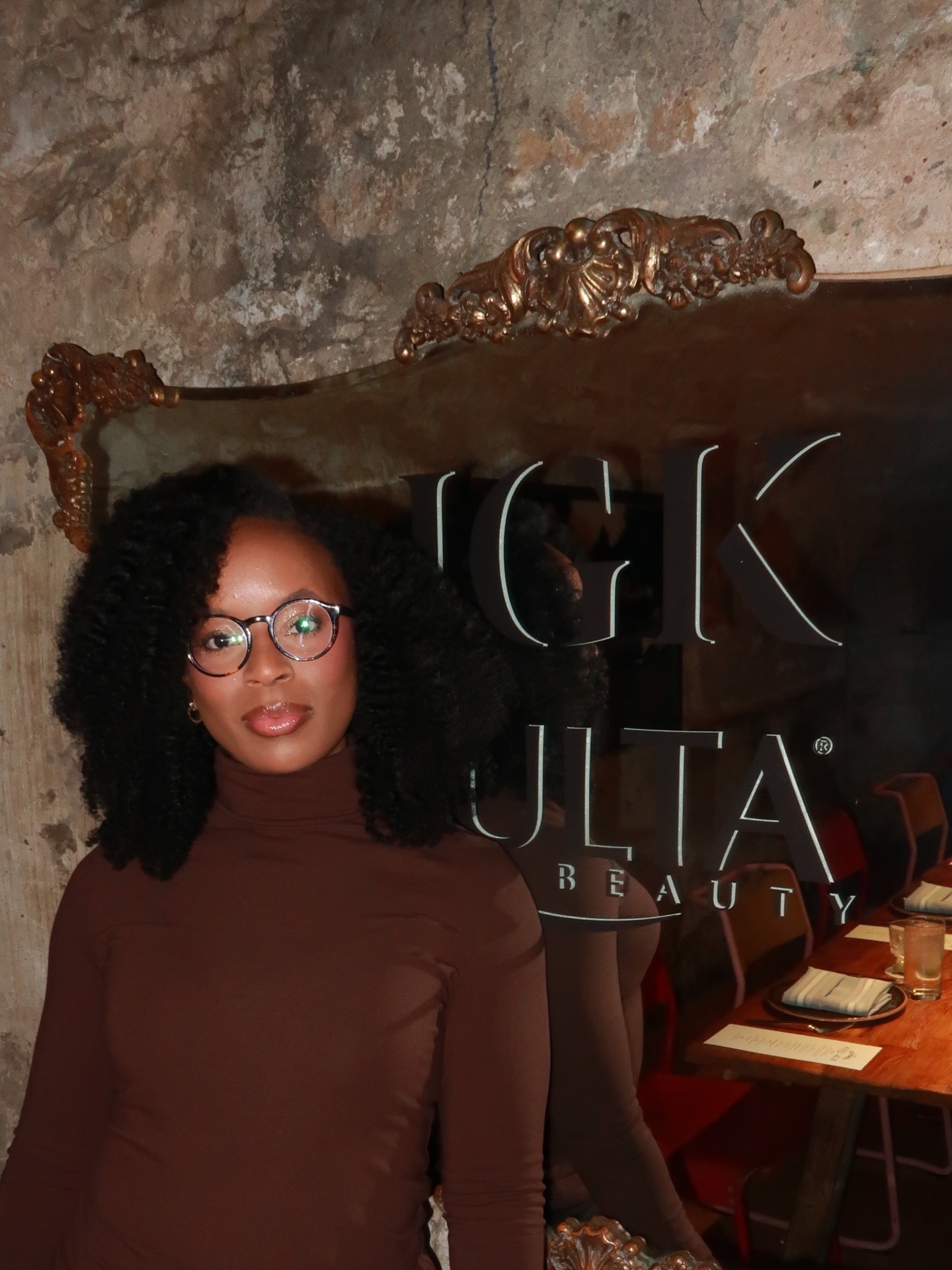 Woman with curly hair and glasses sitting indoors, background includes a decorative wooden frame and a sign with partially visible text.