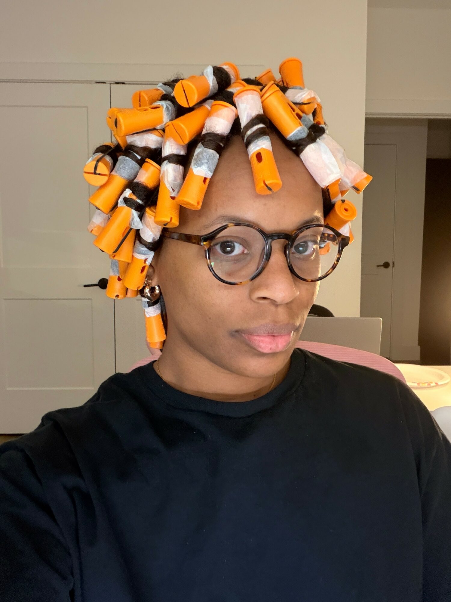 Person with hair styled using orange and black curlers, wearing glasses and a black shirt, indoors.