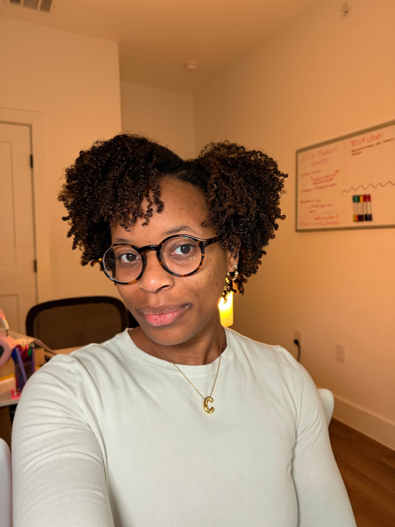 Person with curly hair and glasses taking a selfie in a room with a white wall and a door.