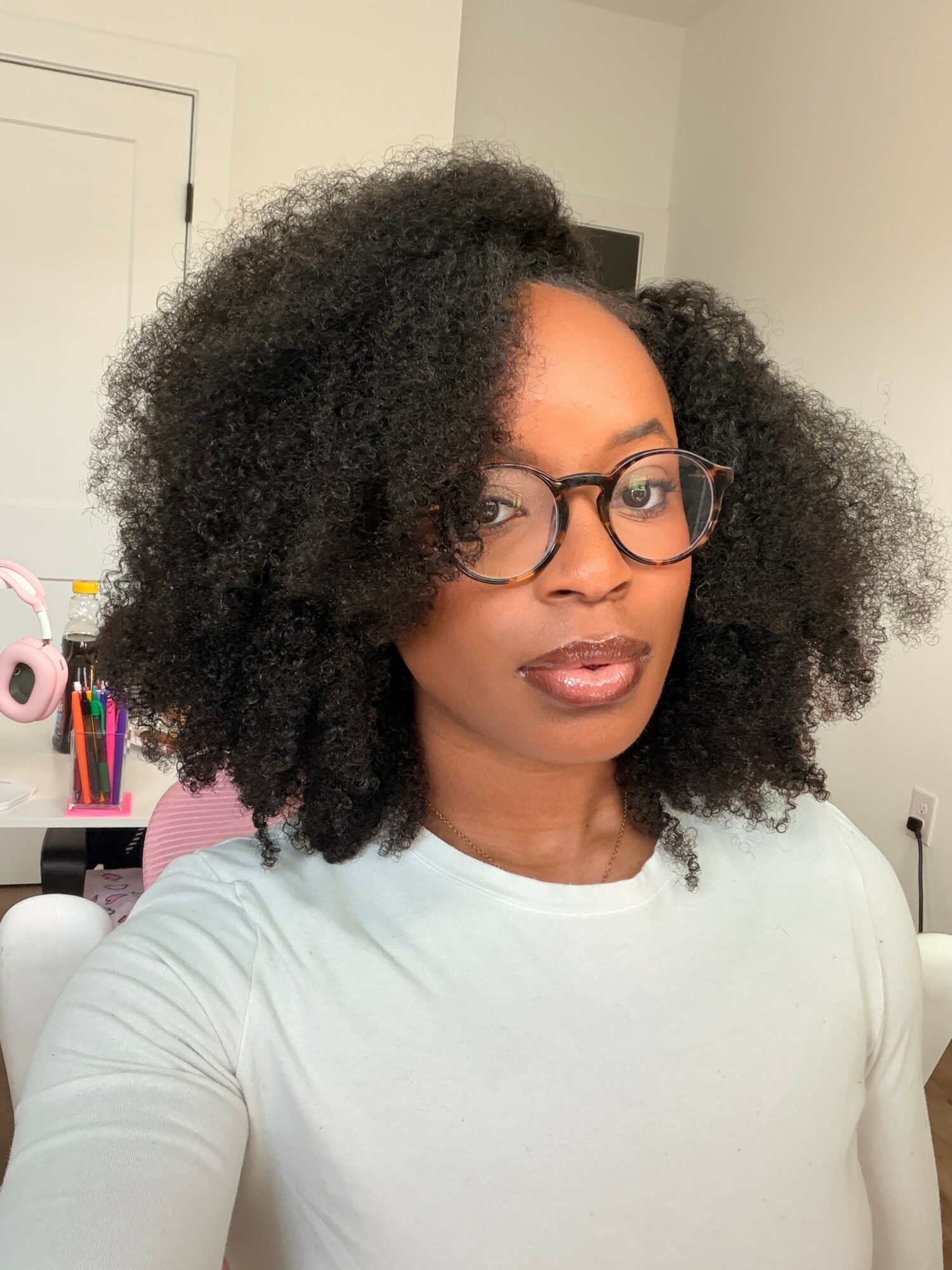 Woman with glasses and natural curly hair taking a selfie indoors, wearing a light-colored top.