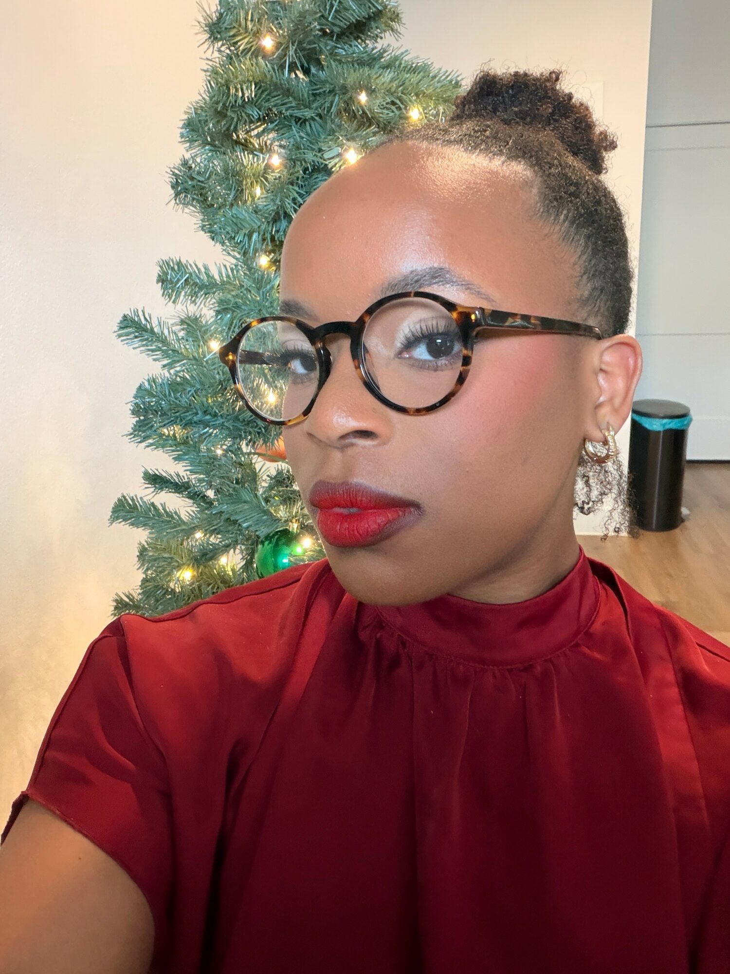Woman with glasses and red lipstick in front of a decorated Christmas tree, wearing a red top.