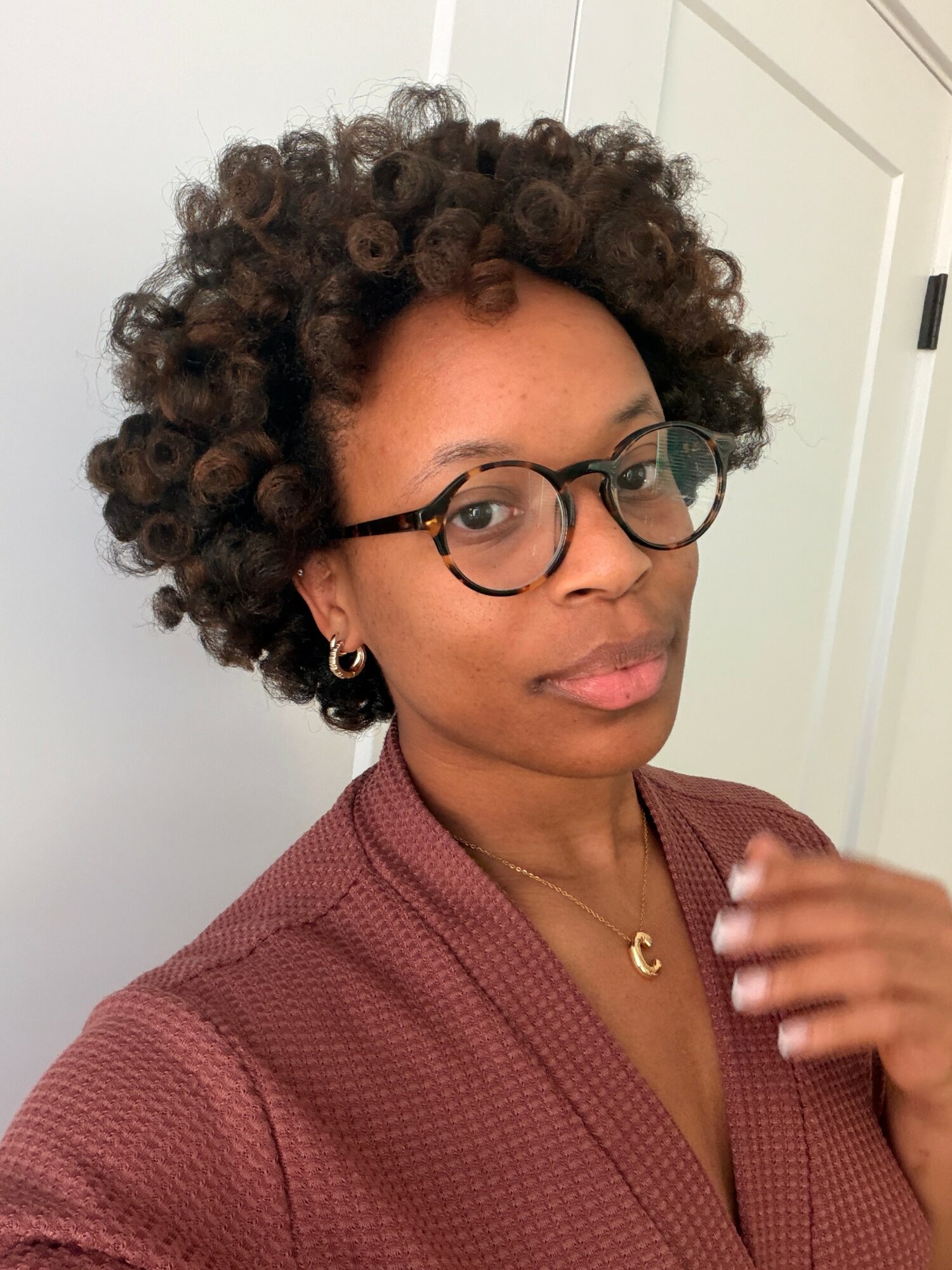 Woman with short curly hair, glasses, and earrings, wearing a maroon top, standing indoors near a door.