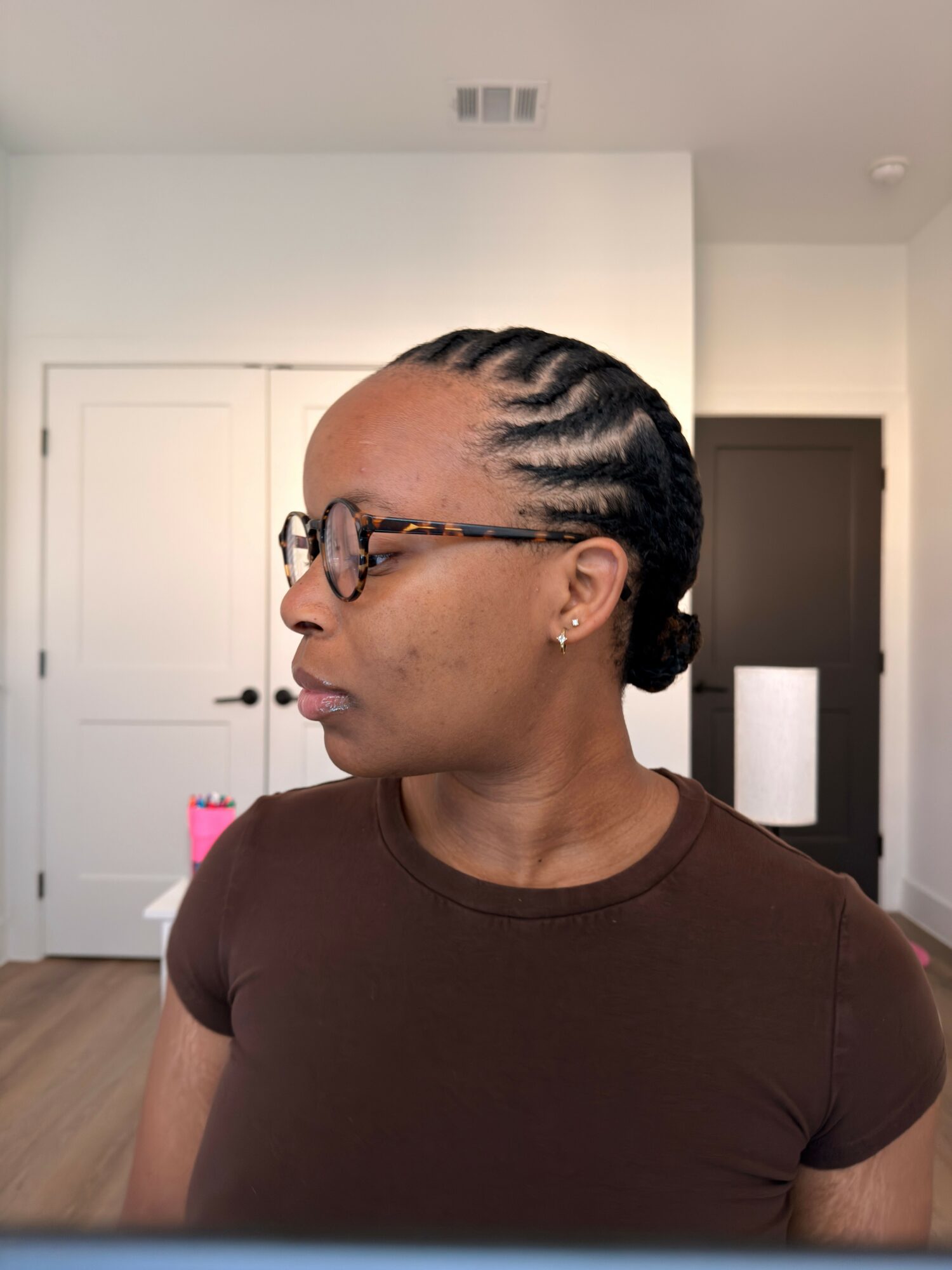 Side view of a woman with braided hair and glasses, wearing a brown shirt, sitting indoors. Background includes a white door and wall.