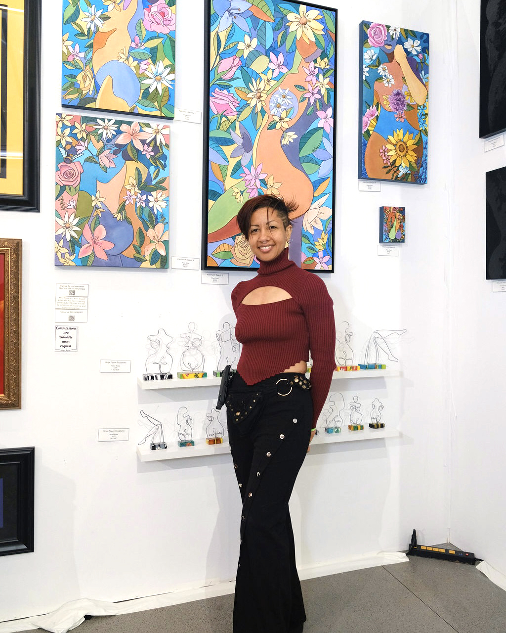 Young woman with short hair smiling in art gallery, colorful paintings of animals and flowers behind her.