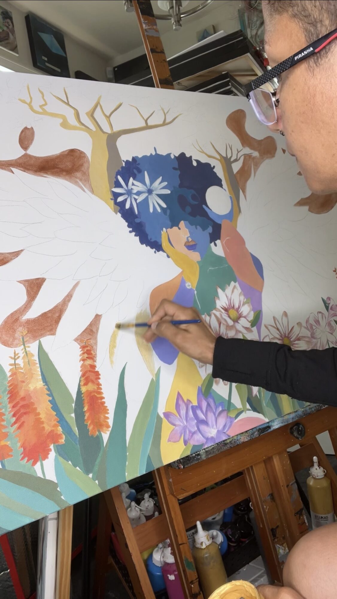Person painting a colorful mural with trees, flowers, and a figure with wings, in an indoor setting.