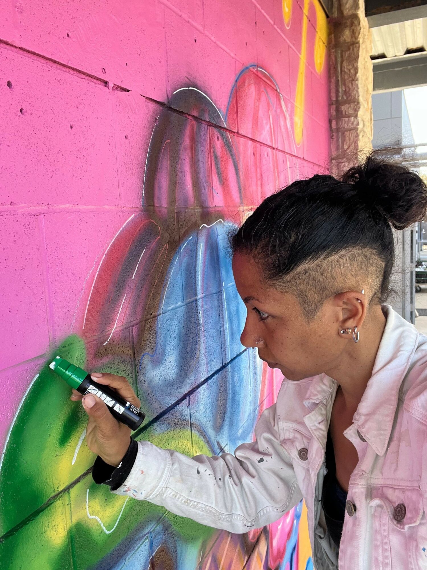 Person drawing colorful graffiti on pink wall with spray paint, leaning close to the artwork, with a bun hairstyle.