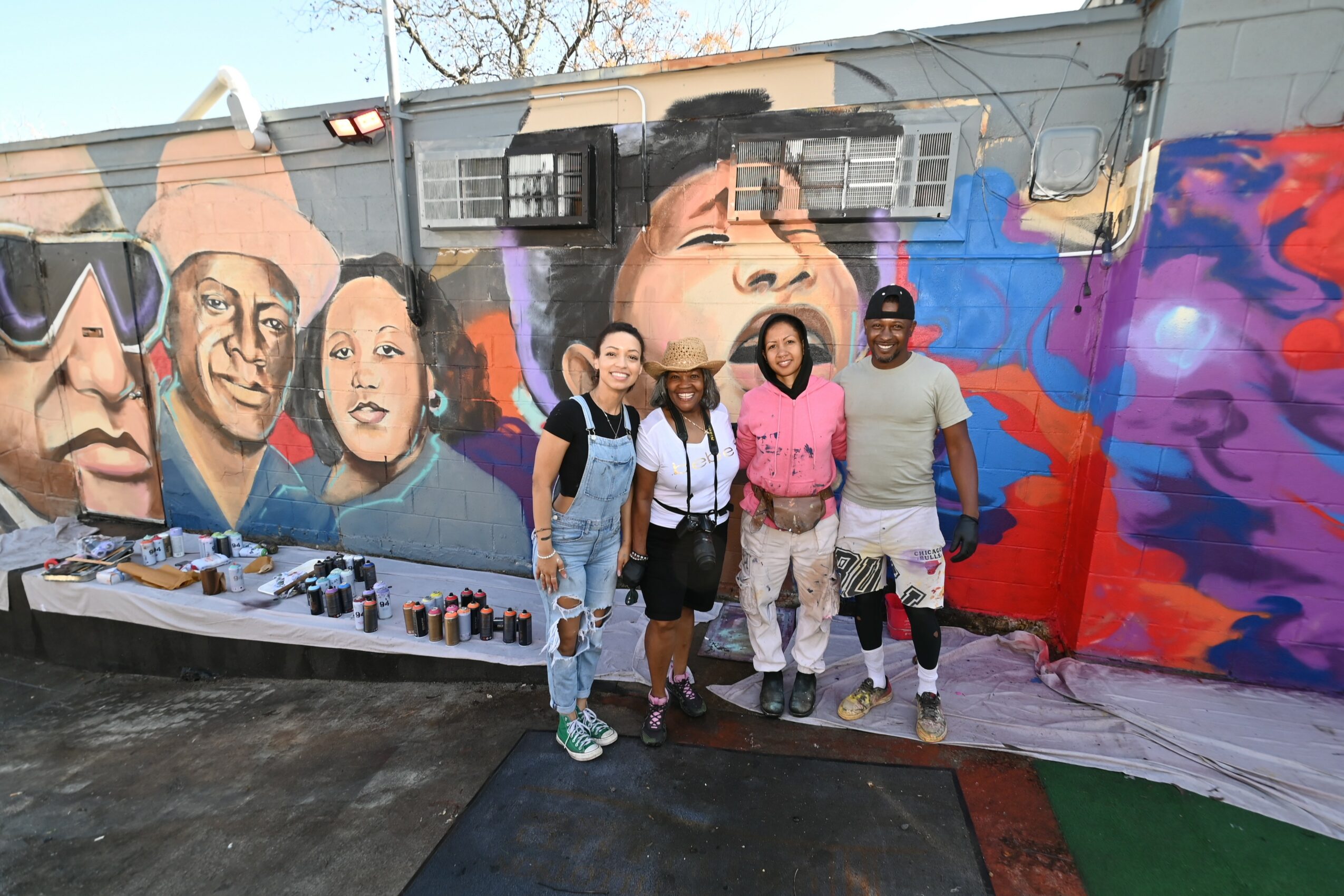 Four people stand in front of a colorful mural with faces painted on a wall, some holding bags, outdoors during daytime.