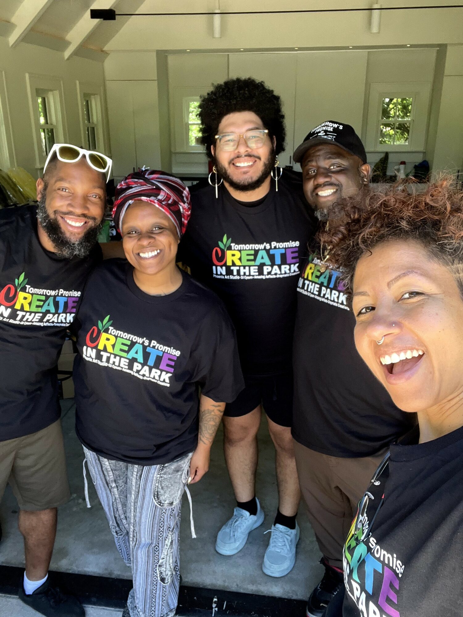 Group of five smiling people wearing matching black T-shirts with colorful text, indoors with windows in the background.