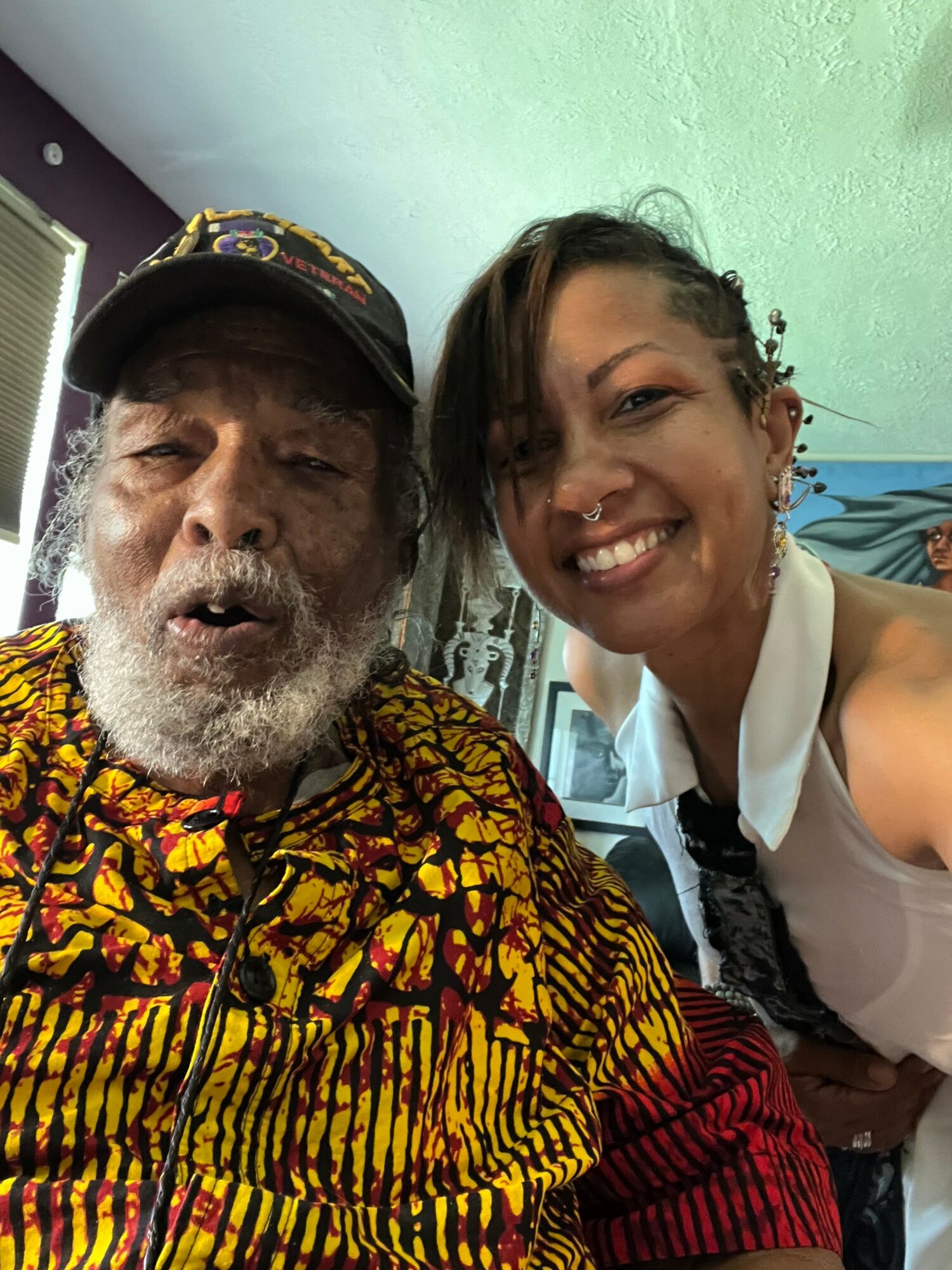 A smiling woman with short hair and a nose piercing takes a selfie with an older man with a beard wearing a colorful shirt and cap.