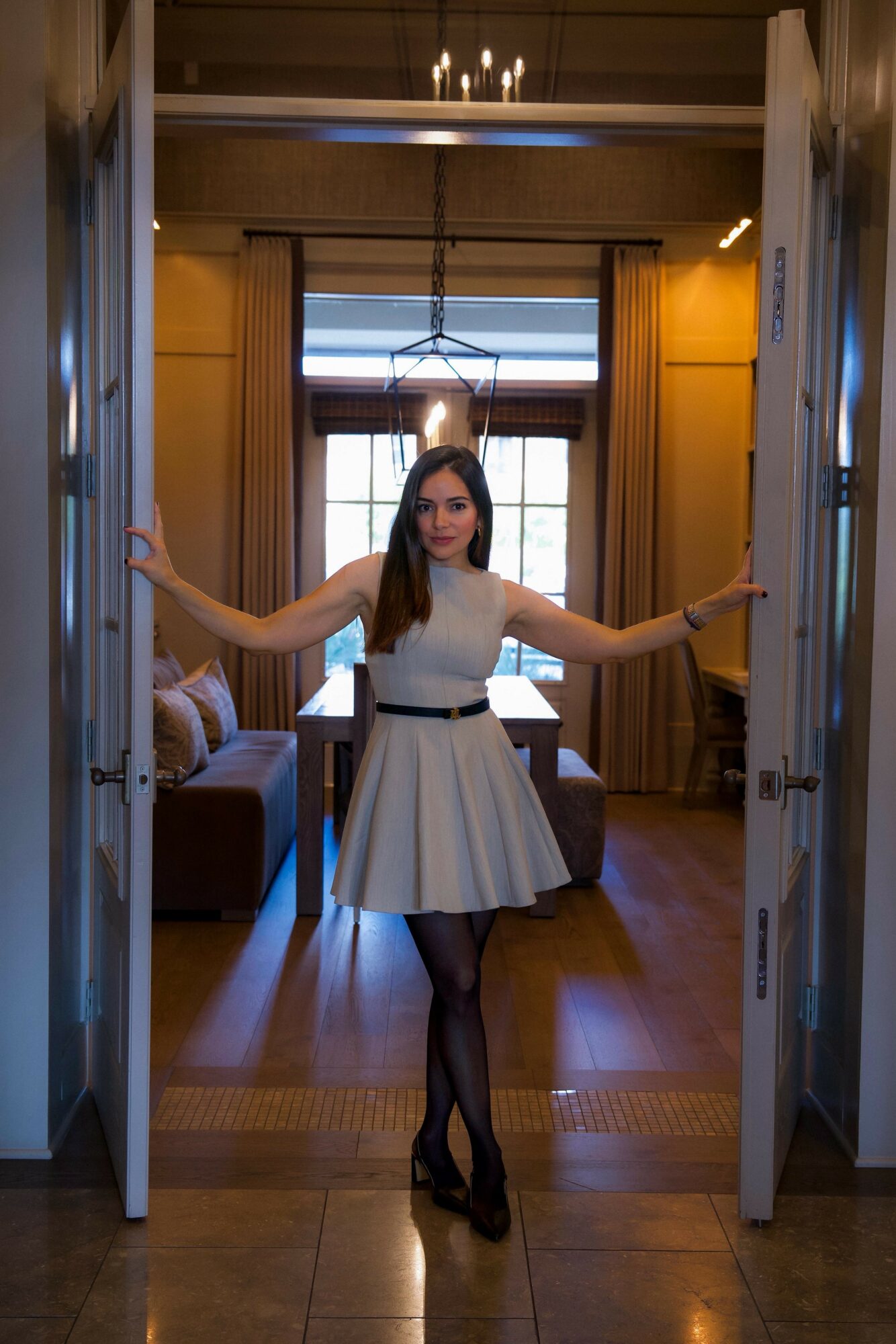 Woman in white dress with black belt standing in doorway with arms outstretched, room with yellow walls and window behind her