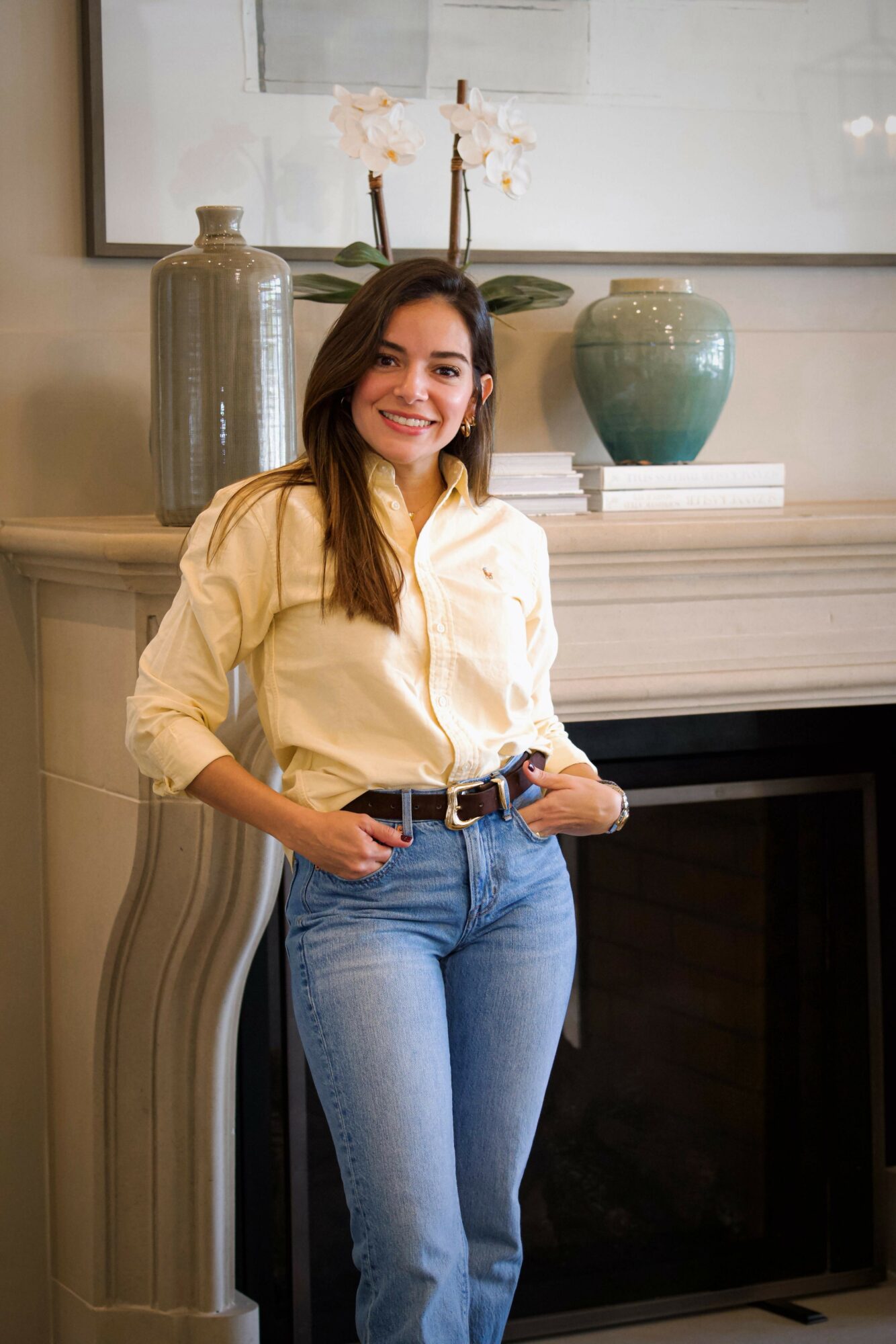 Young woman with long dark hair smiling, standing indoors near a fireplace with decorative vases and a framed picture above.