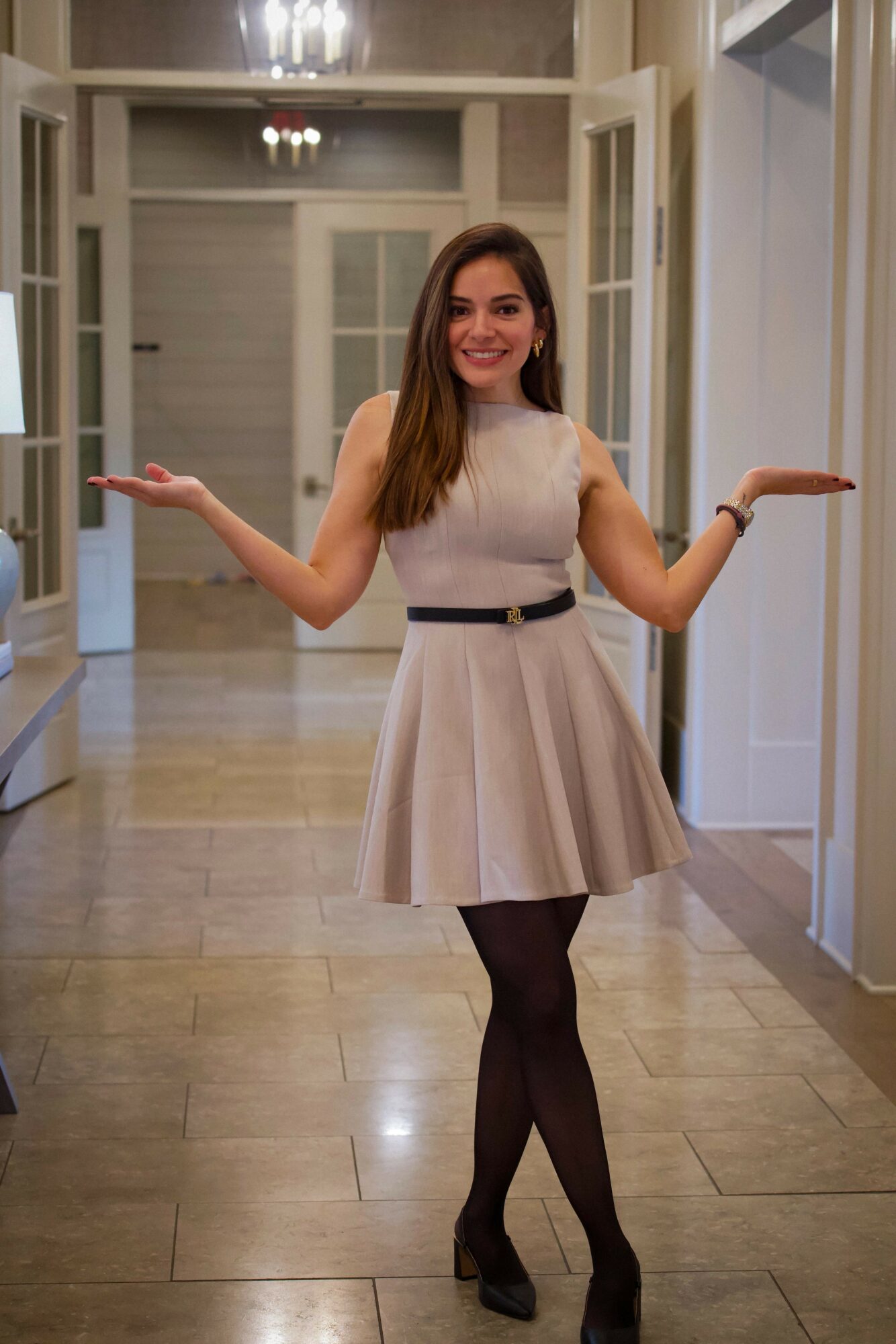 Young woman in a beige dress with a black belt, standing in a hallway with open arms, smiling.