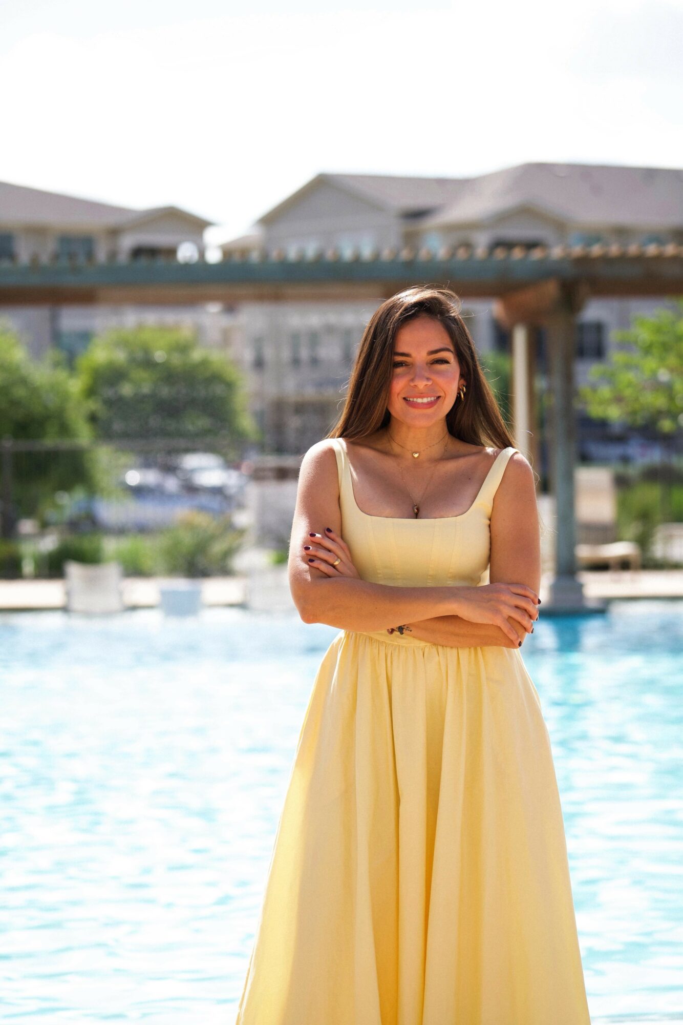Young woman in yellow dress standing by a pool with arms crossed, smiling, buildings in background.
