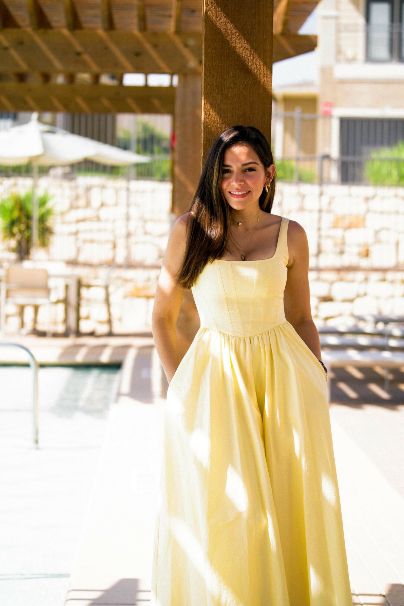 Young woman in yellow dress standing outdoors with a pool and stone wall in background.
