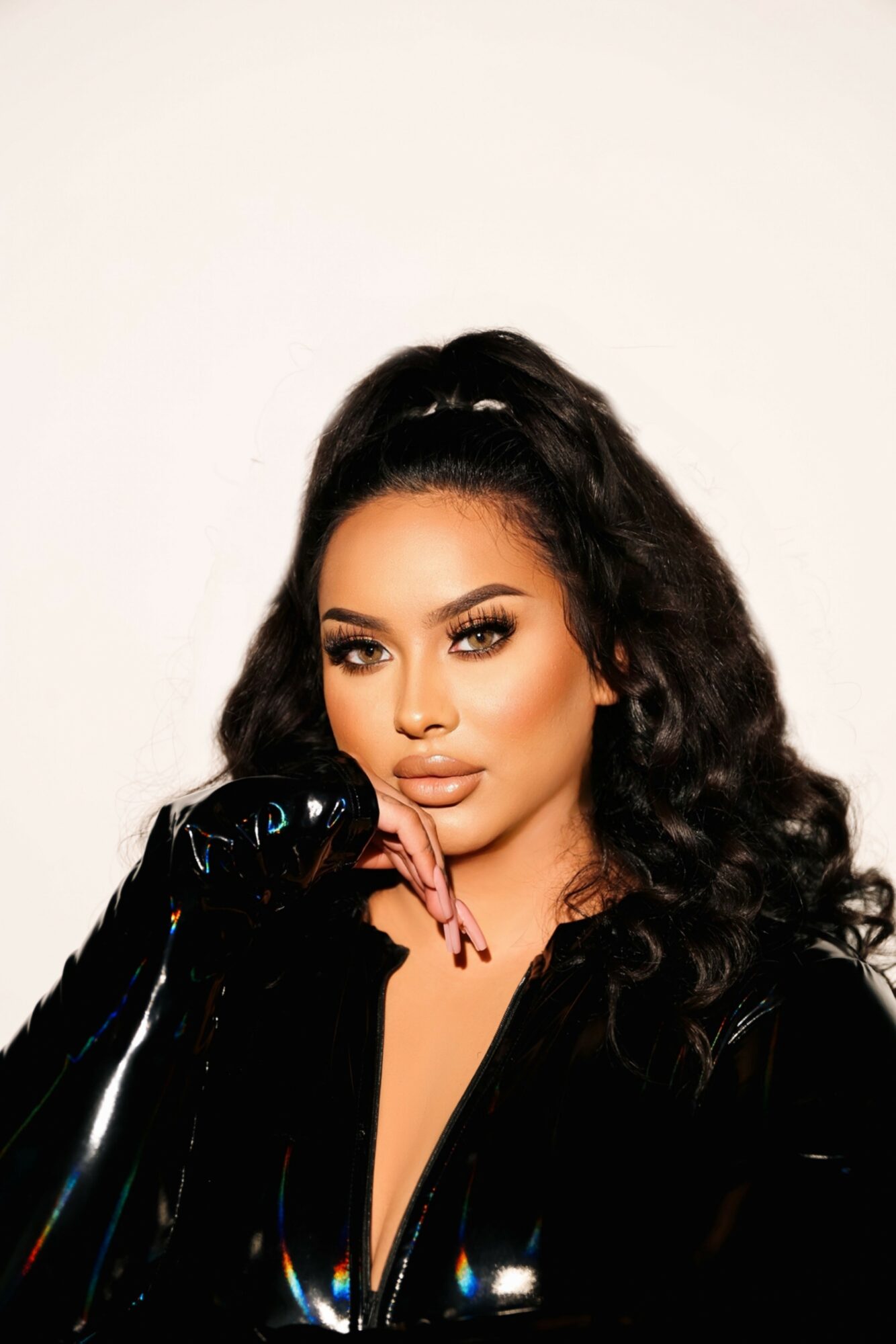 Woman with long, curly hair and makeup, wearing a shiny black outfit, posing with her hand near her face.