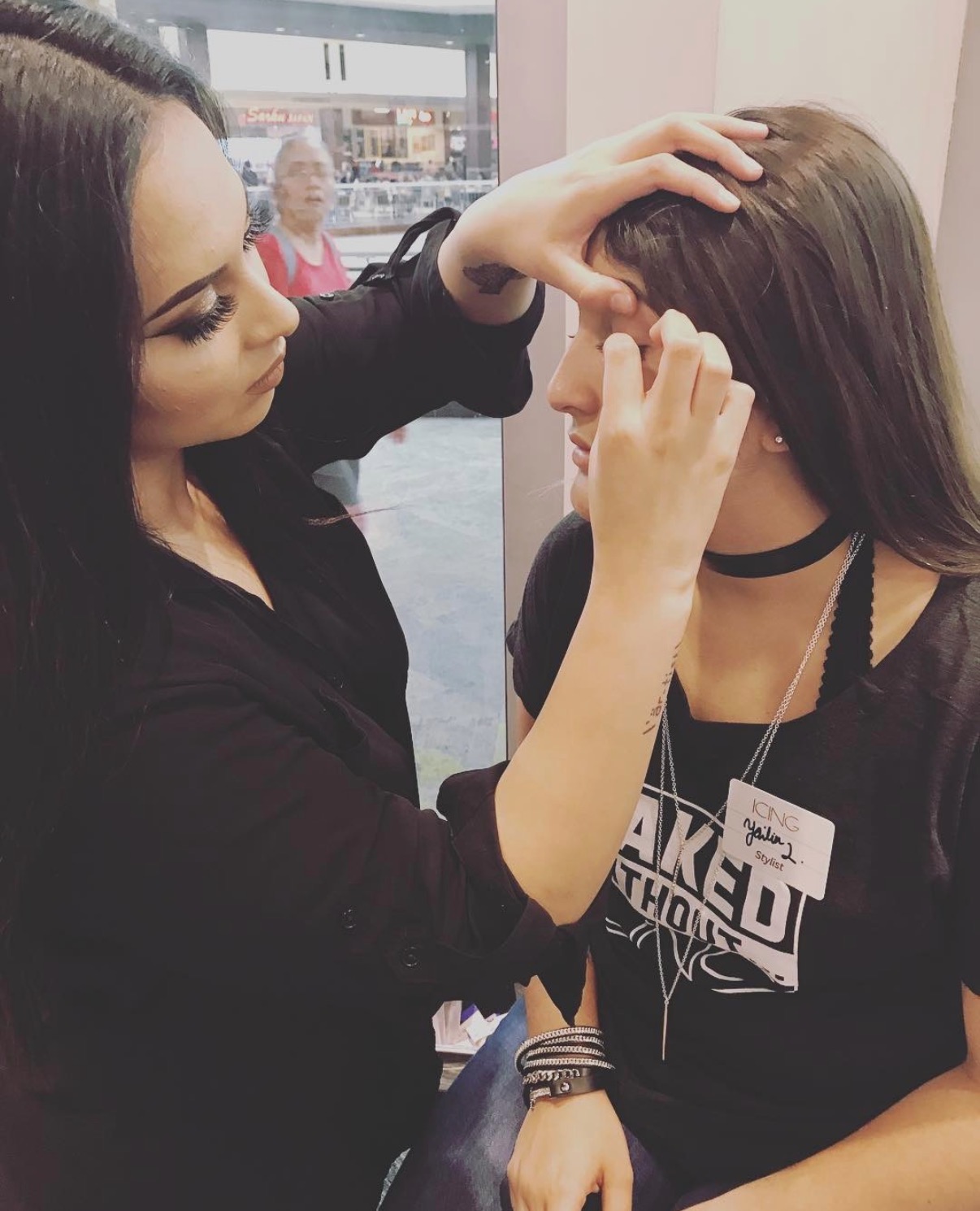 A woman with dark hair applies makeup to a young woman with long brown hair, wearing a black shirt and choker.