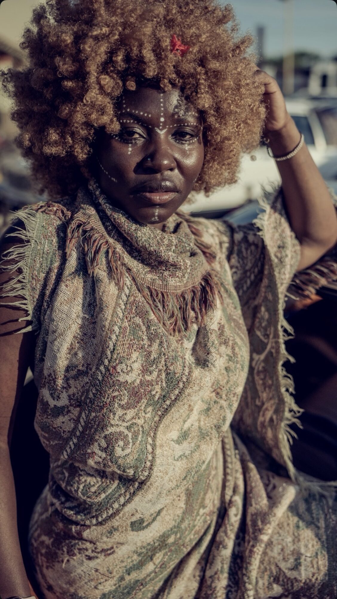 Woman with curly hair and face jewelry, wearing patterned clothing, outdoors with cars in background.