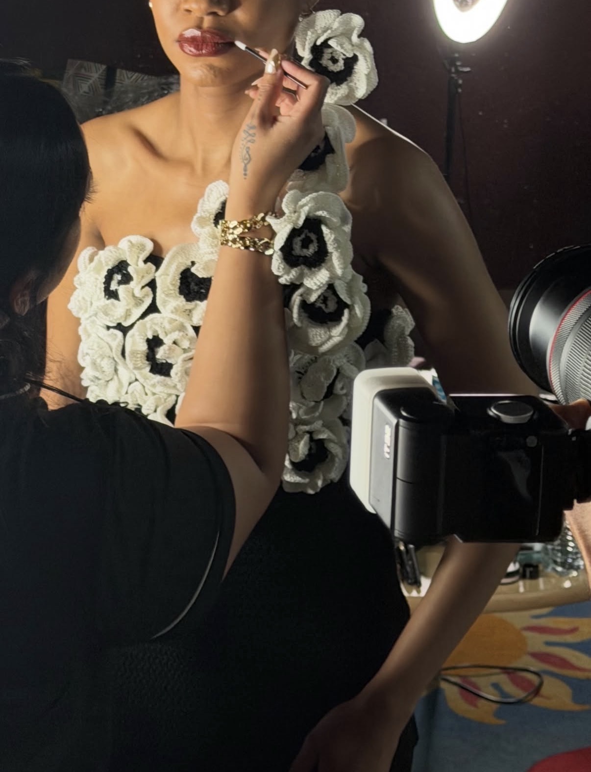Person applying makeup to a woman wearing a floral dress, with a camera and lighting equipment nearby.