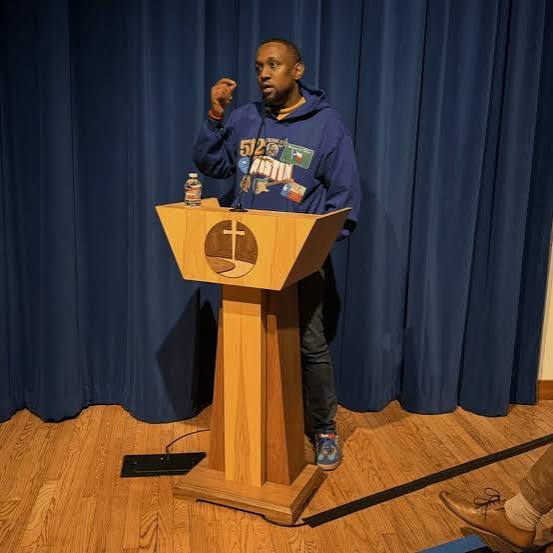 Person standing behind a wooden podium with a blue curtain background, speaking or presenting, with a water bottle on the podium.