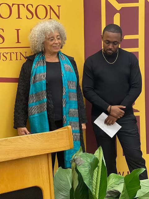 An elderly woman with curly gray hair and a young man with short dark hair stand indoors, with a yellow and purple background and green plants in front.