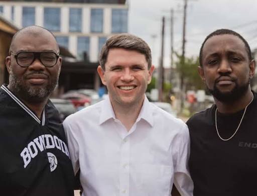 Three men standing outdoors, smiling, with buildings and power lines in the background.