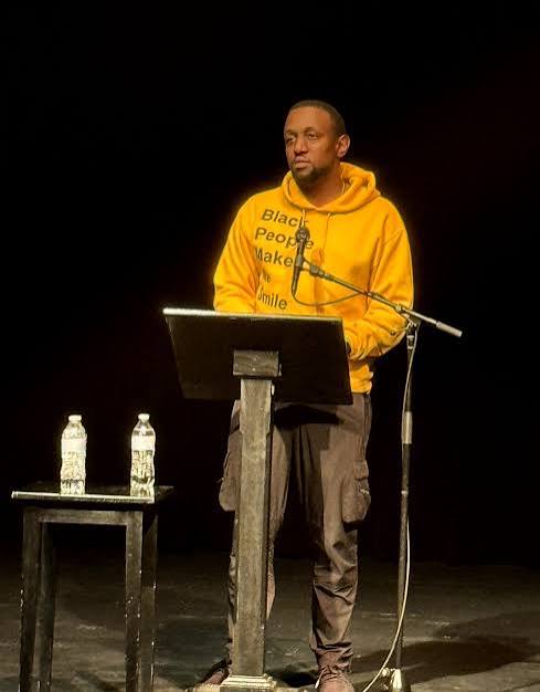 Man in yellow hoodie speaking at a podium with two water bottles on a table, on a dark stage.