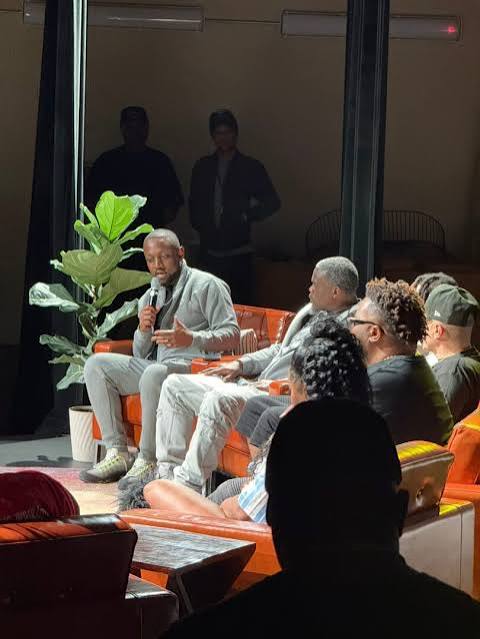 Three men seated on a stage, one speaking into a microphone, with an audience in front, and a large plant behind them.