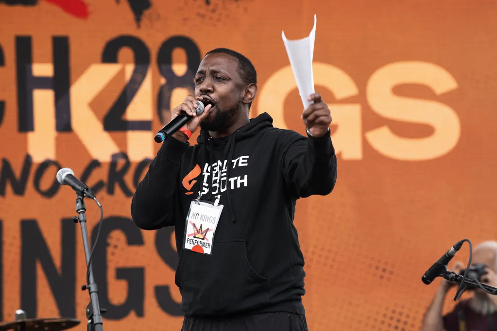 A man speaking into a microphone on stage, holding papers, with a large orange background and text behind him.