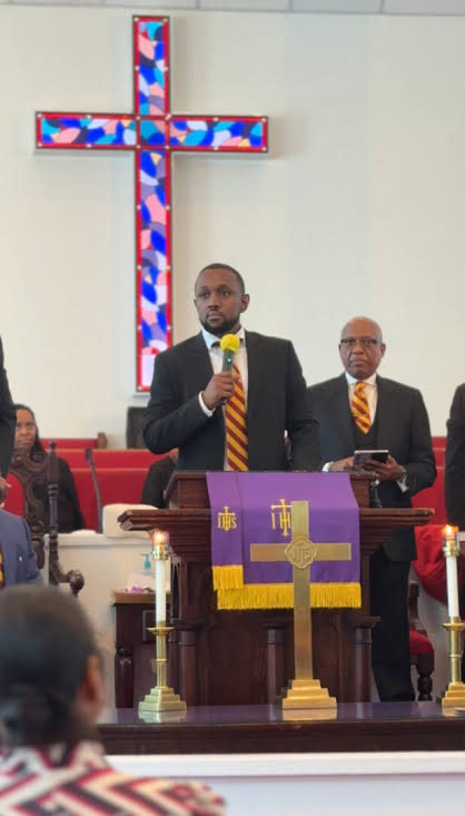 Man in suit and yellow tie speaking at church pulpit with cross and congregation present.