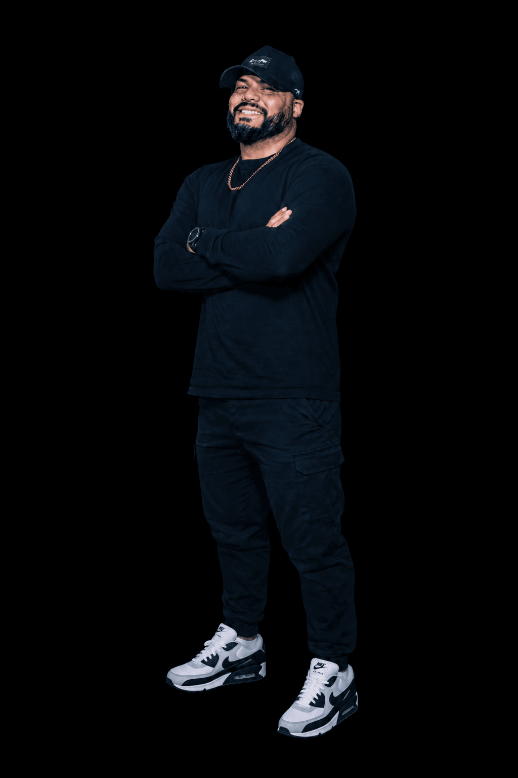Man with a beard smiling, wearing a black cap, black long-sleeve shirt, black pants, and white sneakers, standing with arms crossed.