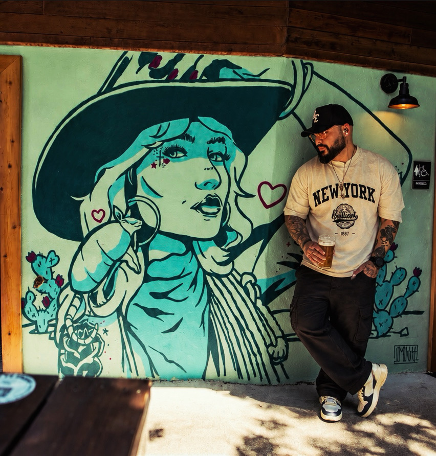 Man with tattoos and glasses leaning against a wall with a large mural of a woman in a hat, holding a phone, with hearts around her.