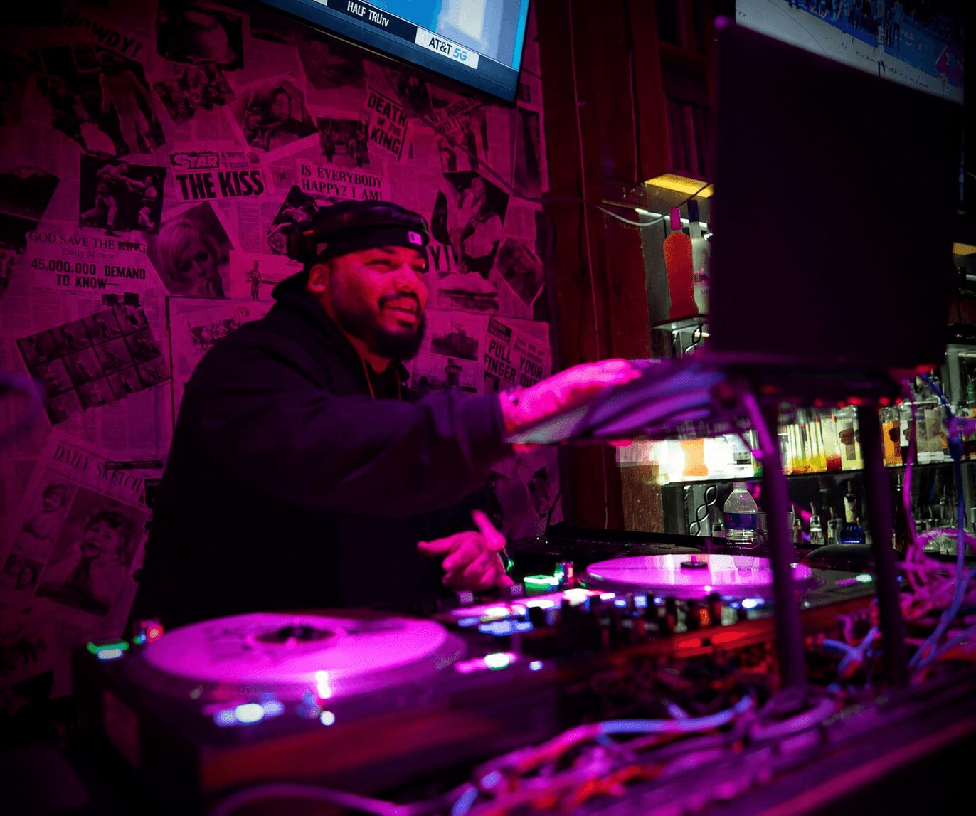 DJ smiling and mixing music at a nightclub with pink lighting and newspaper-themed wallpaper, equipment in foreground.