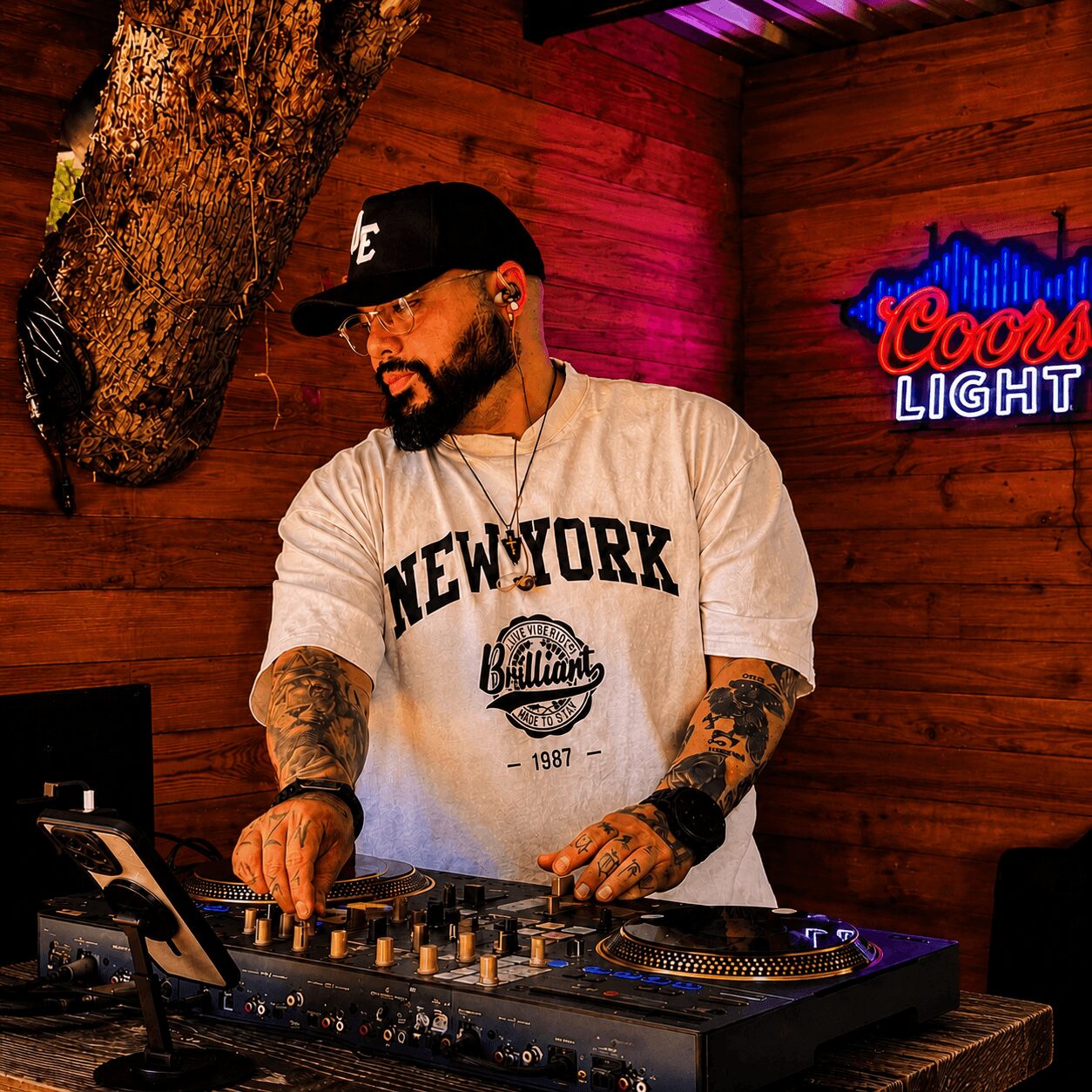 Man with tattoos and beard wearing a white 'New York' t-shirt, DJing in a wooden-paneled room with neon sign.