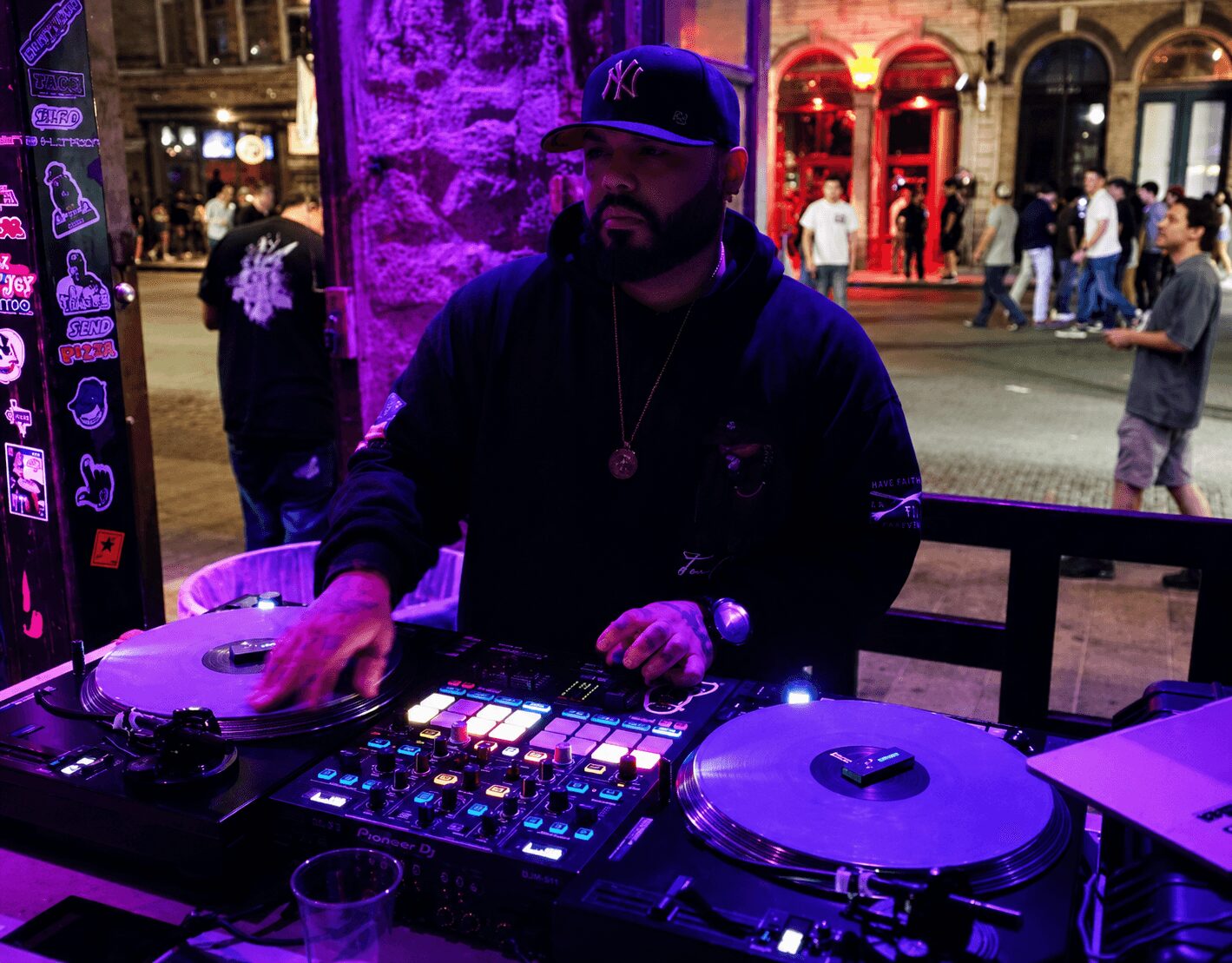DJ wearing a cap and hoodie, mixing music at an outdoor event at night, with people walking in the background.