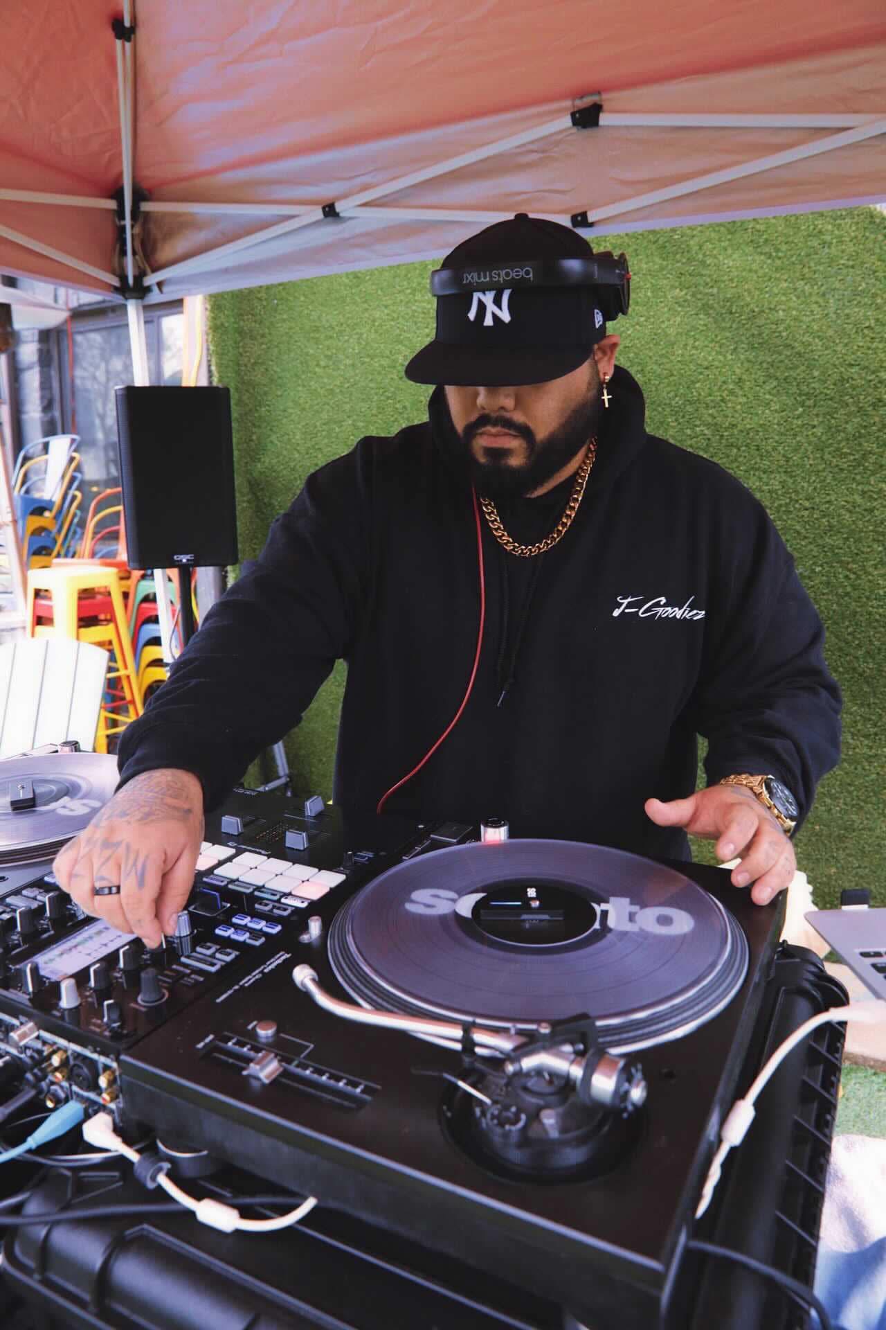 DJ wearing a black cap and hoodie, operating turntables and mixer outdoors under a canopy.