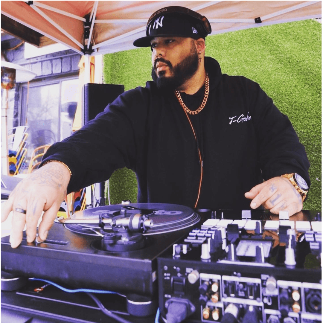 Man DJing with turntable and mixer, wearing a black hoodie, cap, and gold chain, under a tent outdoors.