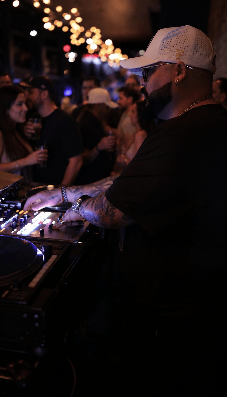 Person wearing a white cap and black shirt DJing at a crowded outdoor event at night, with blurred lights in background.