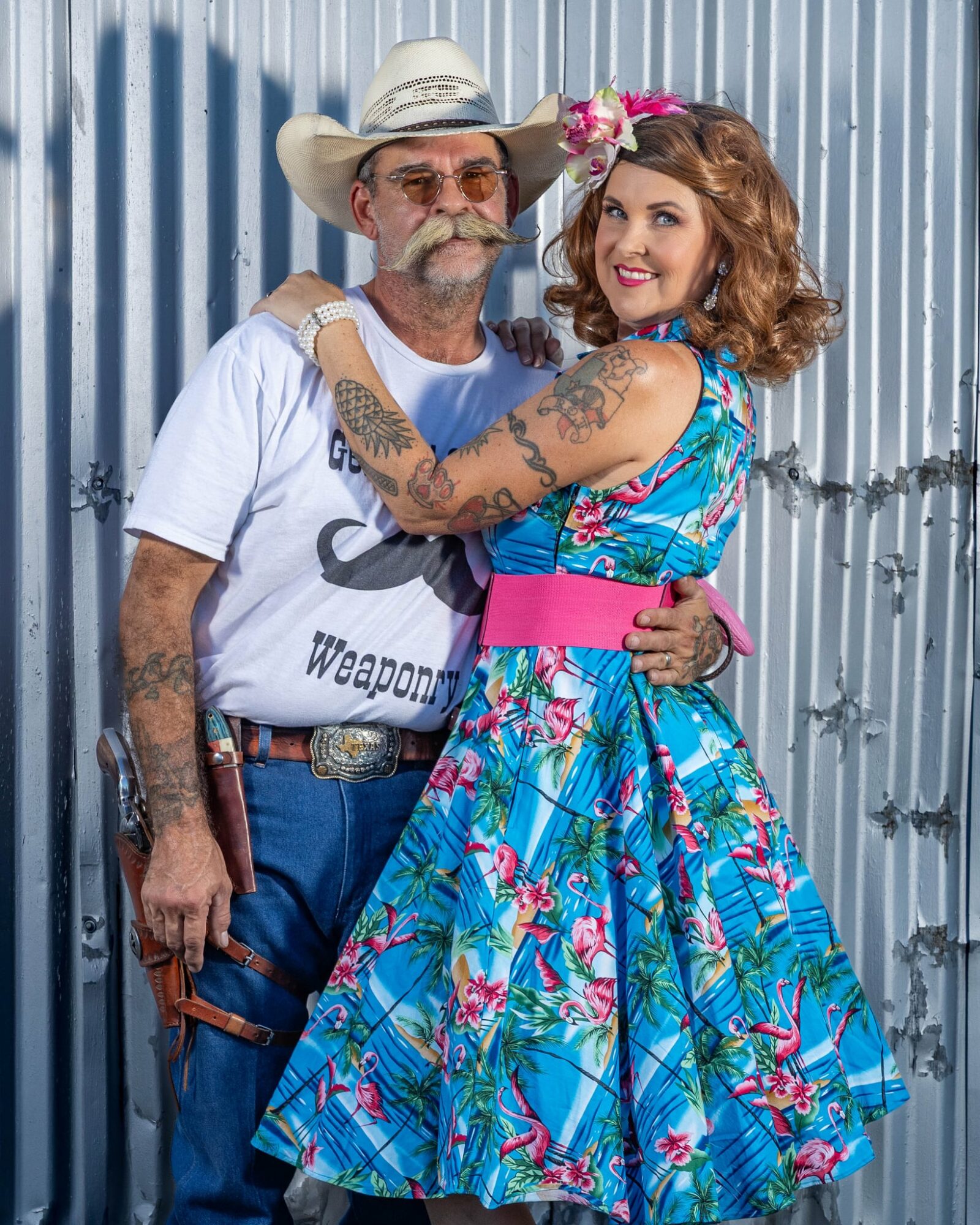 A man and woman stand close together, smiling, with a metal fence background. The woman has floral dress and pink flowers in her hair.