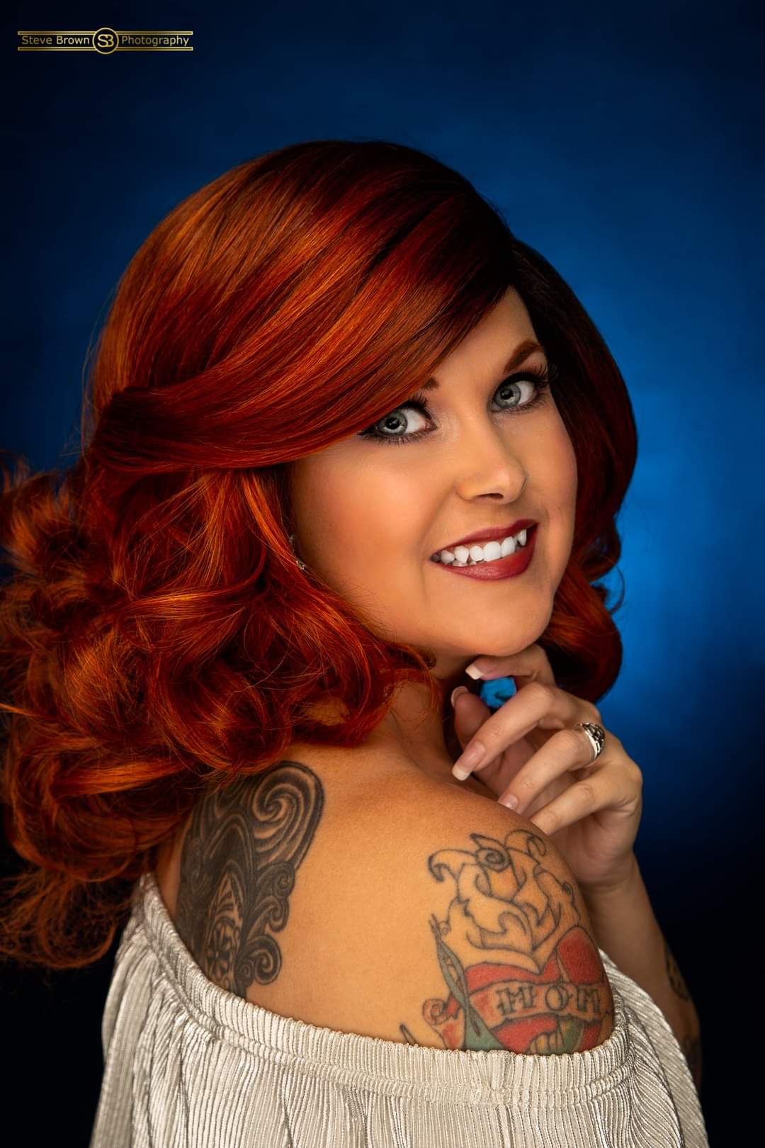Woman with red hair, smiling, showing tattoos on shoulder and arm, wearing a white top, against a blue background.