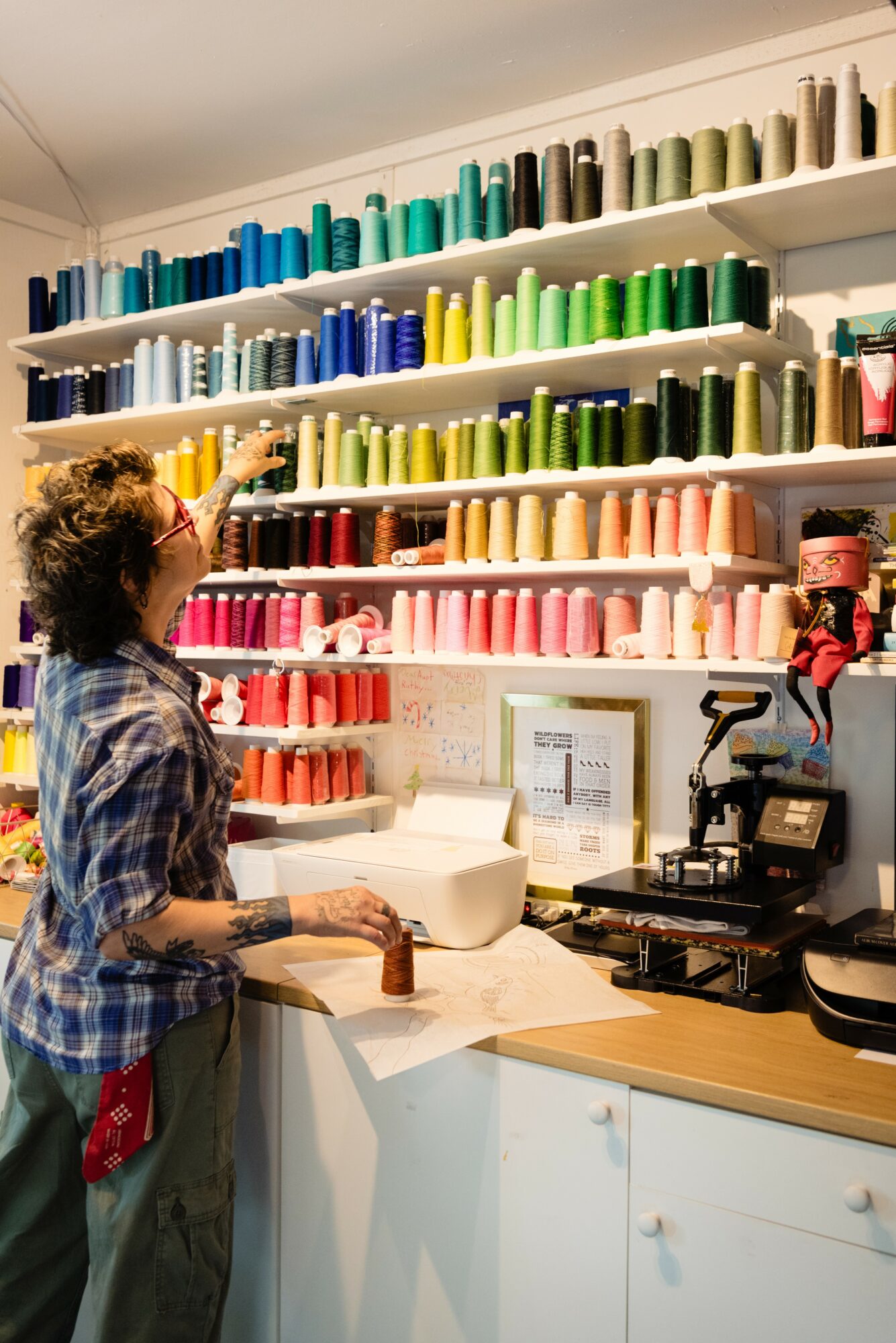 Person selecting thread from colorful spools on wall shelves in a sewing shop, with sewing machine and supplies on counter.