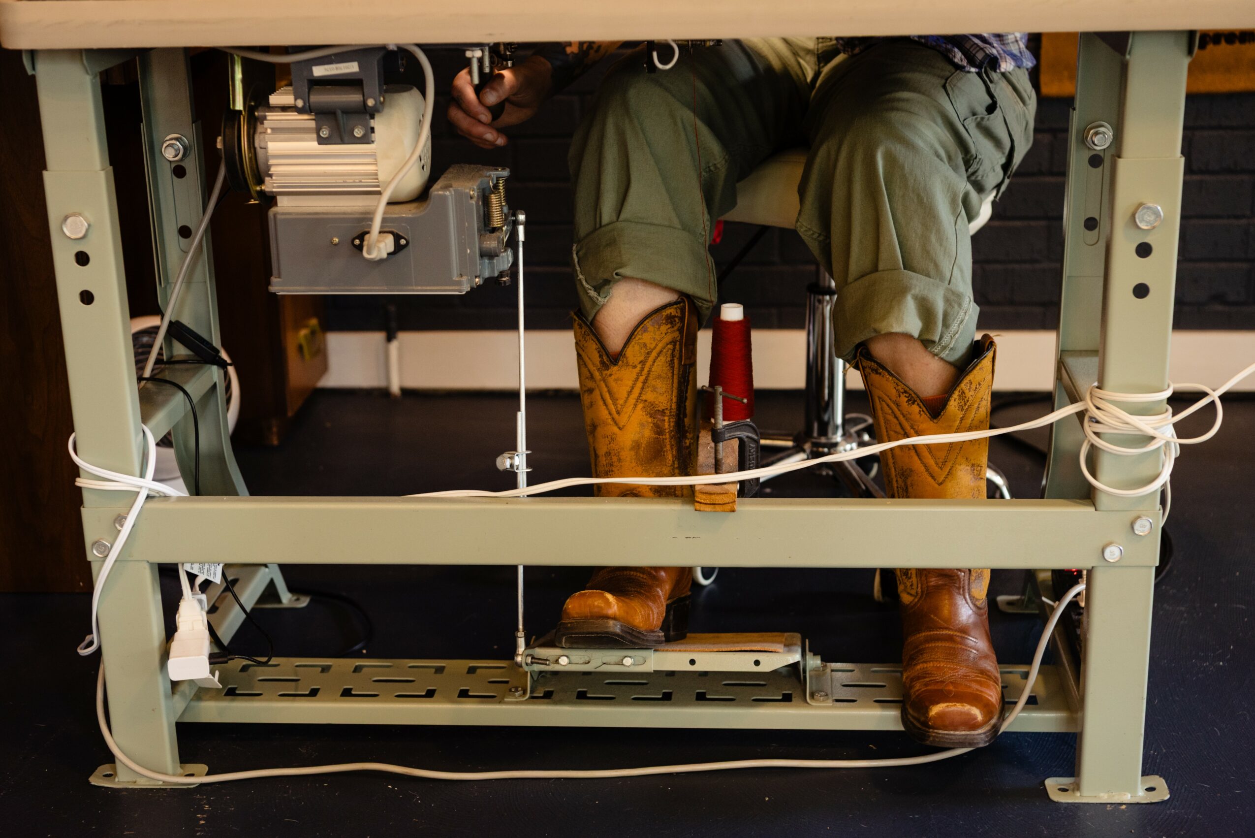 Person wearing green pants and brown boots working at a desk with wires and equipment underneath.