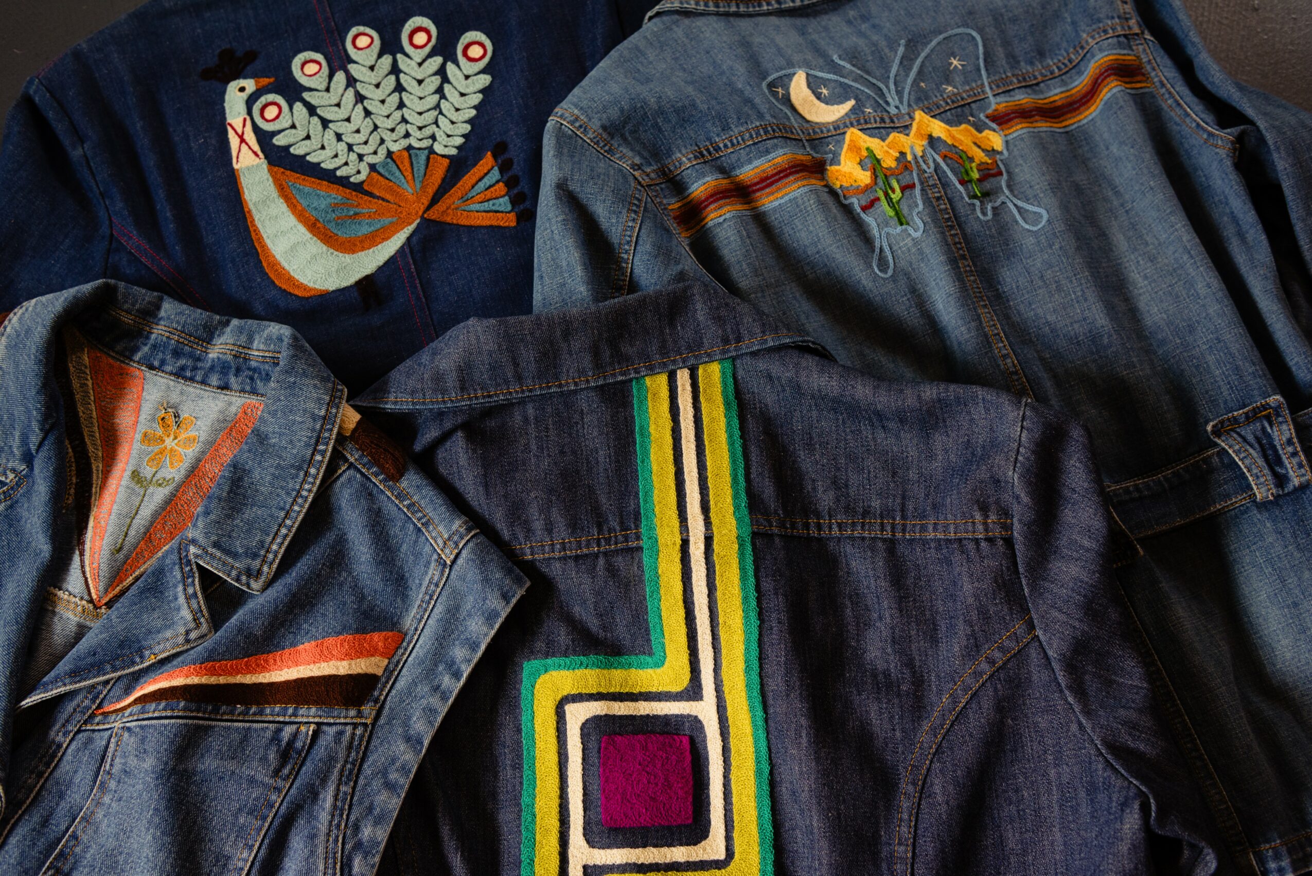 Three denim jackets with colorful embroidered designs on the back, including a peacock, a moon with a dragonfly, and a rainbow stripe pattern.