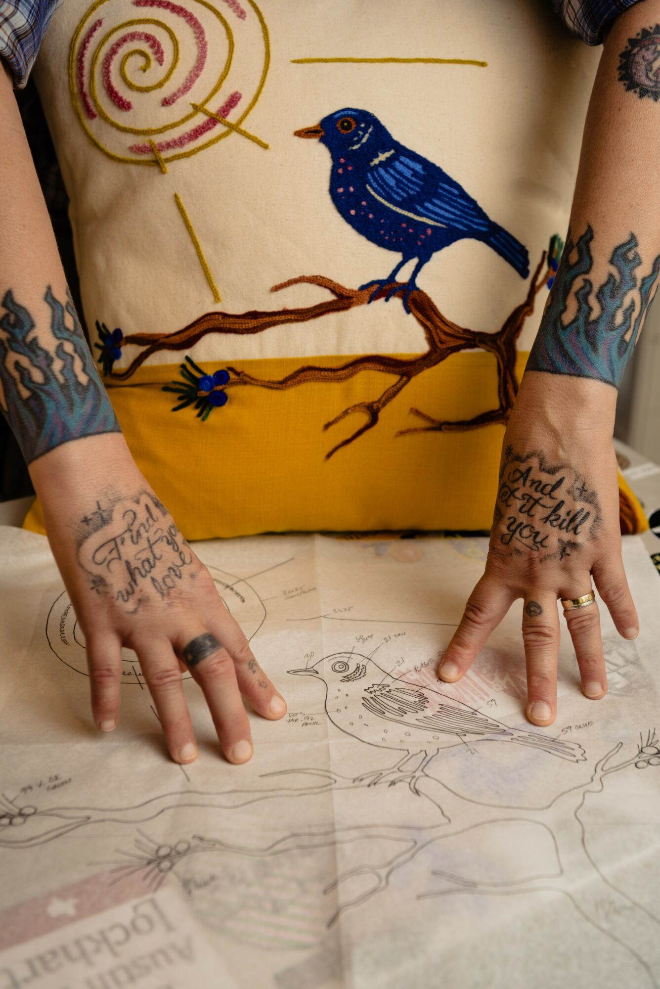 Person with tattoos on hands and arms, drawing on paper with a pencil, colorful body paint of a bird on torso, sun, and branch.